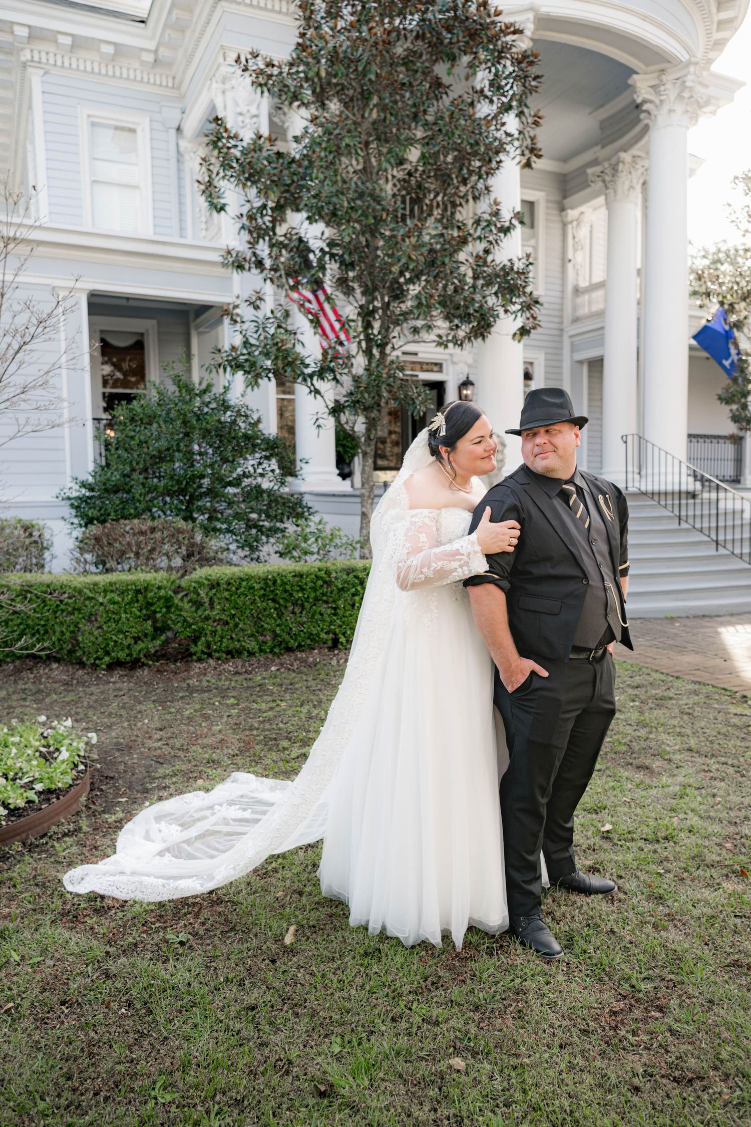 Bride in white lace gown with veil and groom in black suit pose together outside a grand white Southern mansion.