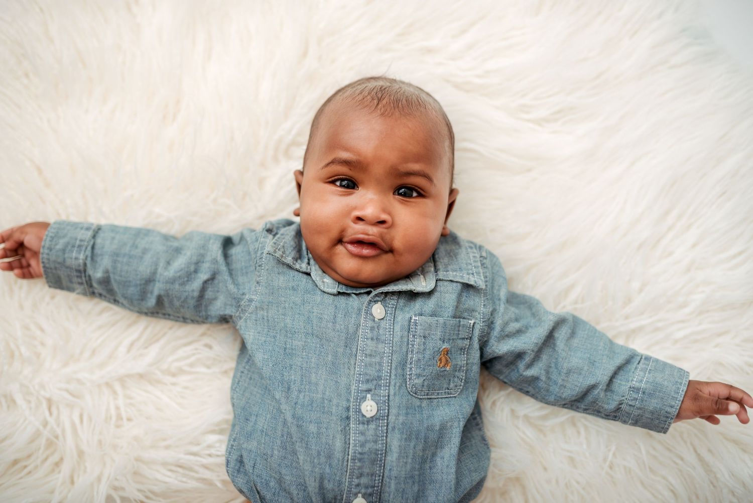 Baby in chambray shirt lying on white fur rug during photo session.