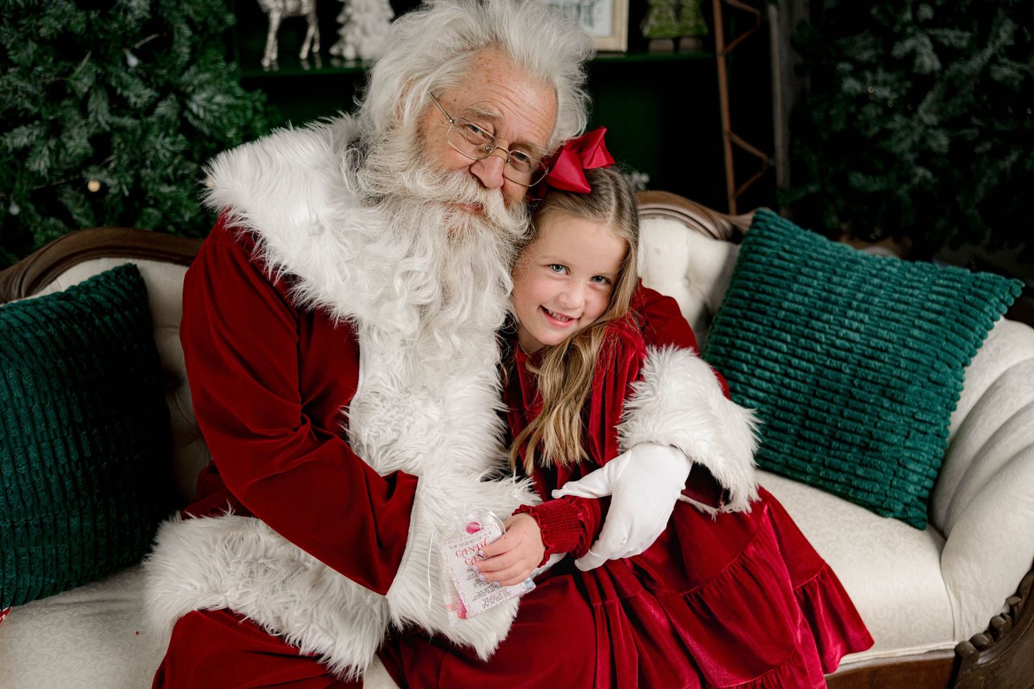 Young girl in red dress sitting on Santa Claus lap, smiling together on a white couch with holiday decor.