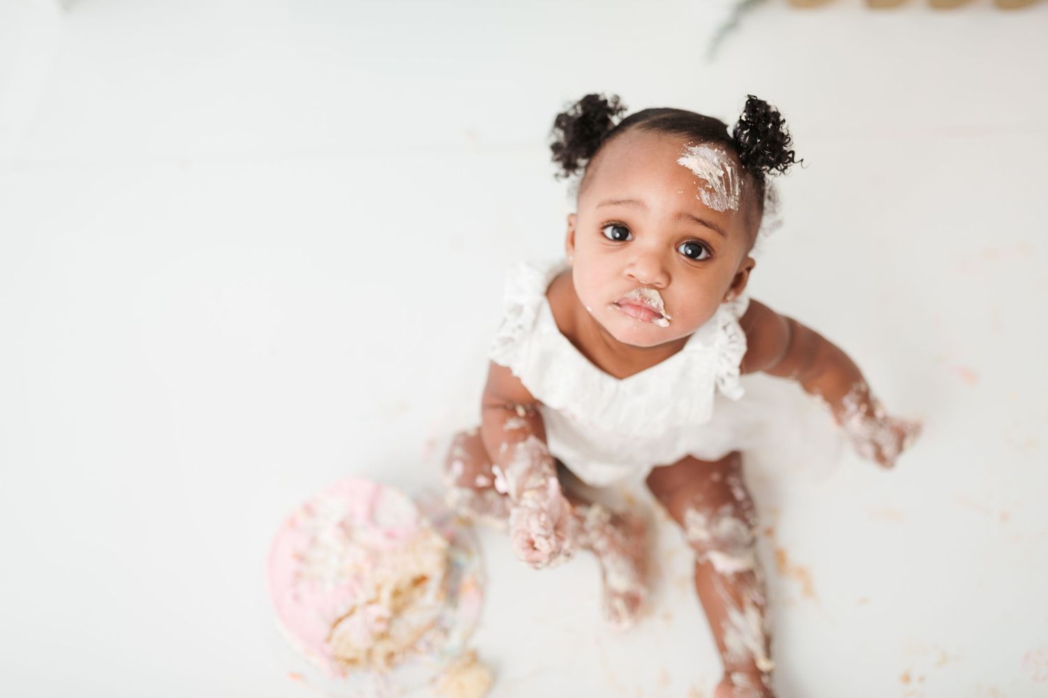 Adorable baby girl in white dress covered in cake frosting during first birthday cake smash photo session.