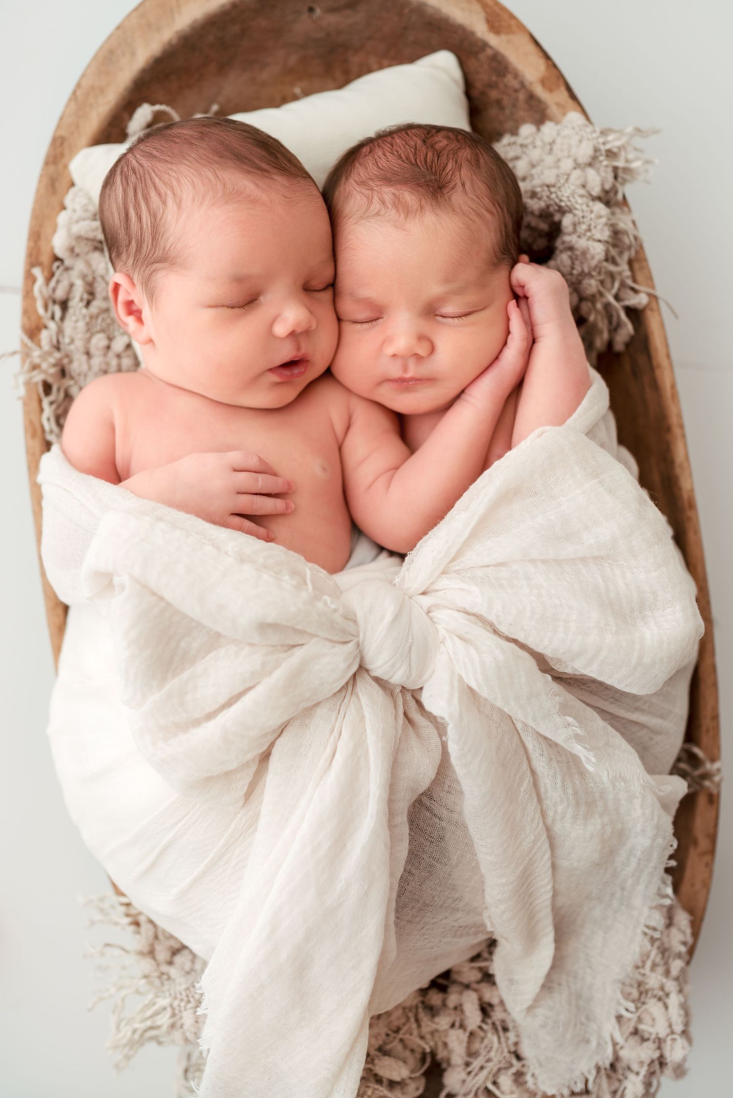 Two sleeping newborn babies swaddled in white blankets nestled together in a cozy brown basket.