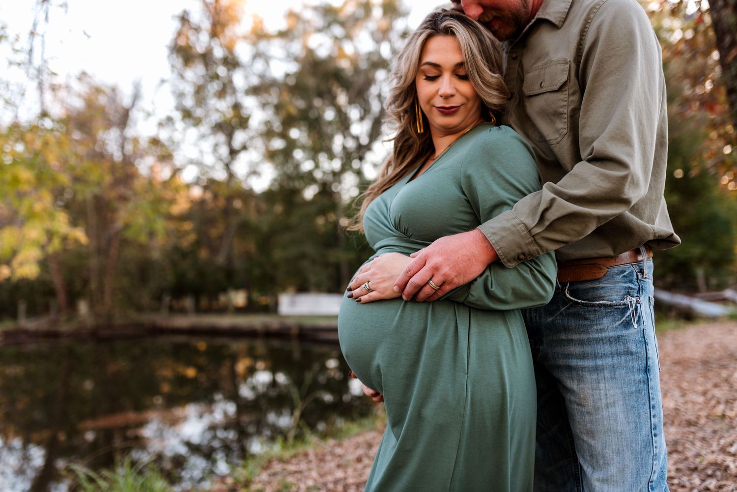 Couple embraces in a tender maternity moment by a pond during golden hour.
