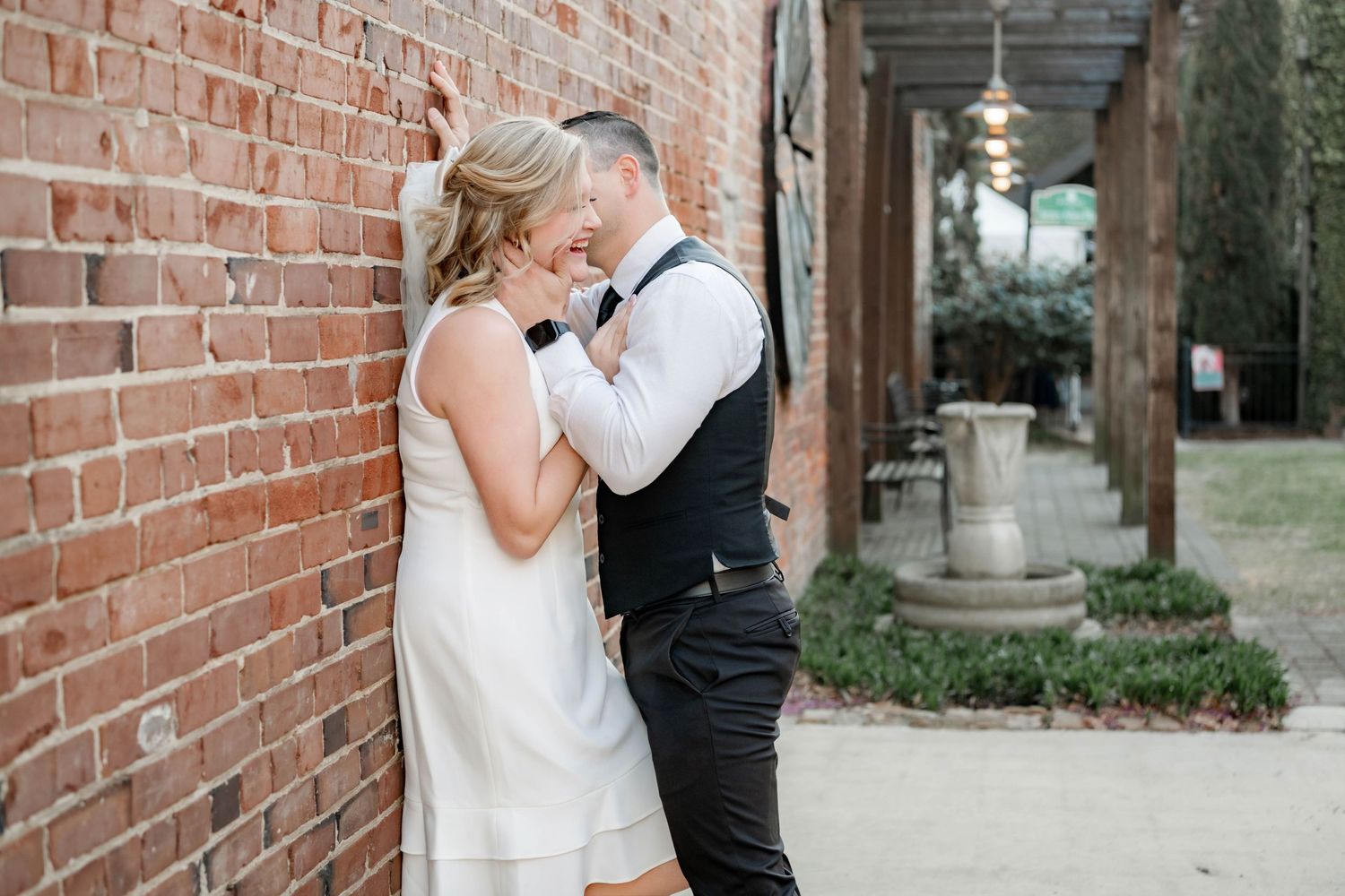 A bride and groom share an intimate moment leaning against a rustic brick wall at their outdoor wedding venue.
