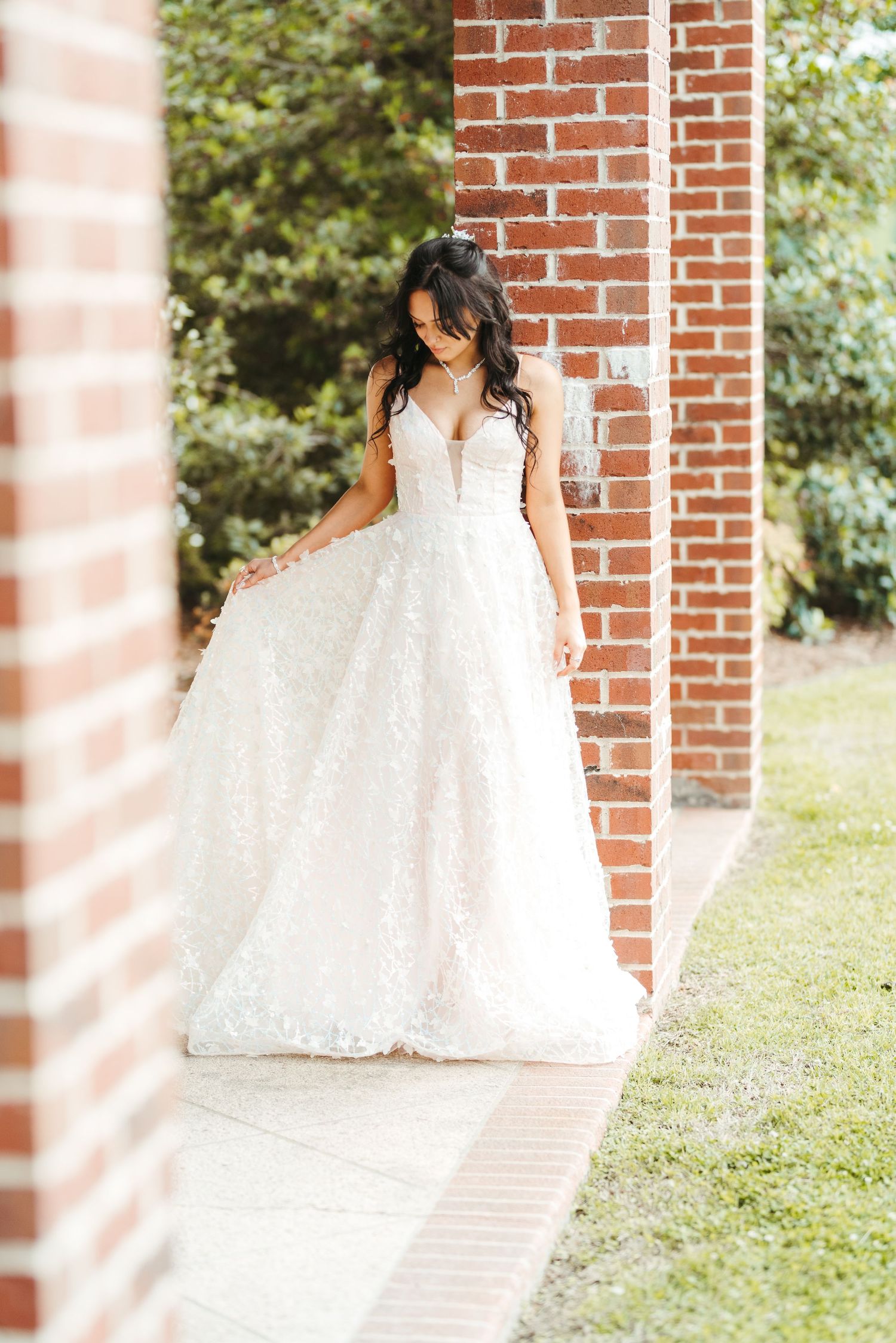 An elegant bride in a white lace ball gown poses between brick columns on a garden pathway.