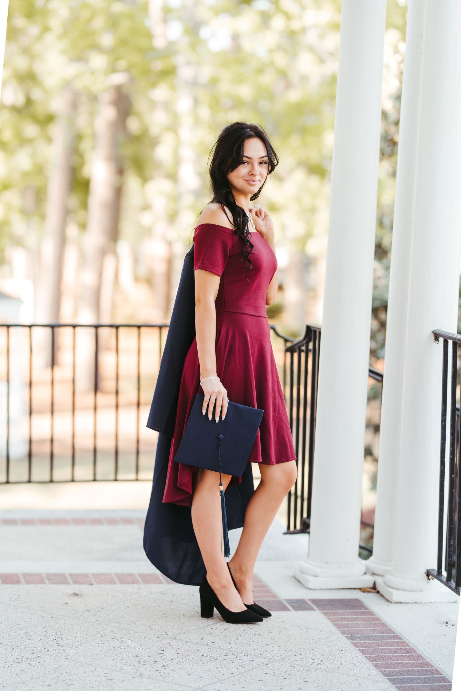 A stylish woman in a burgundy dress and black coat poses by white columns on a porch.