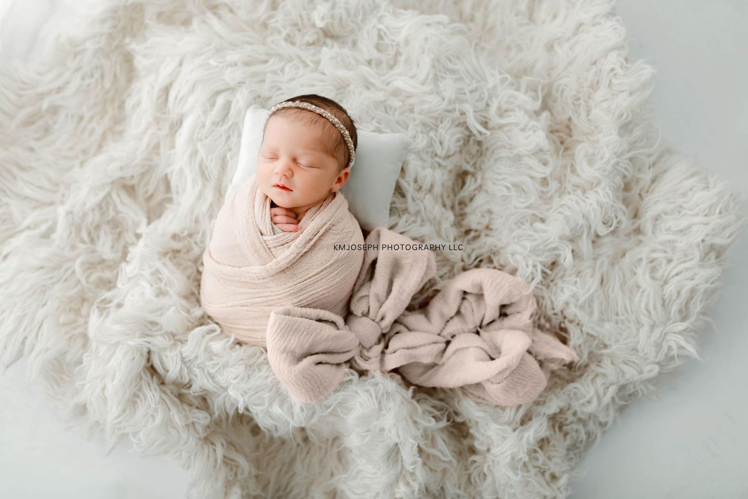 Sleeping newborn wrapped in beige fabric lying on fluffy white blanket.