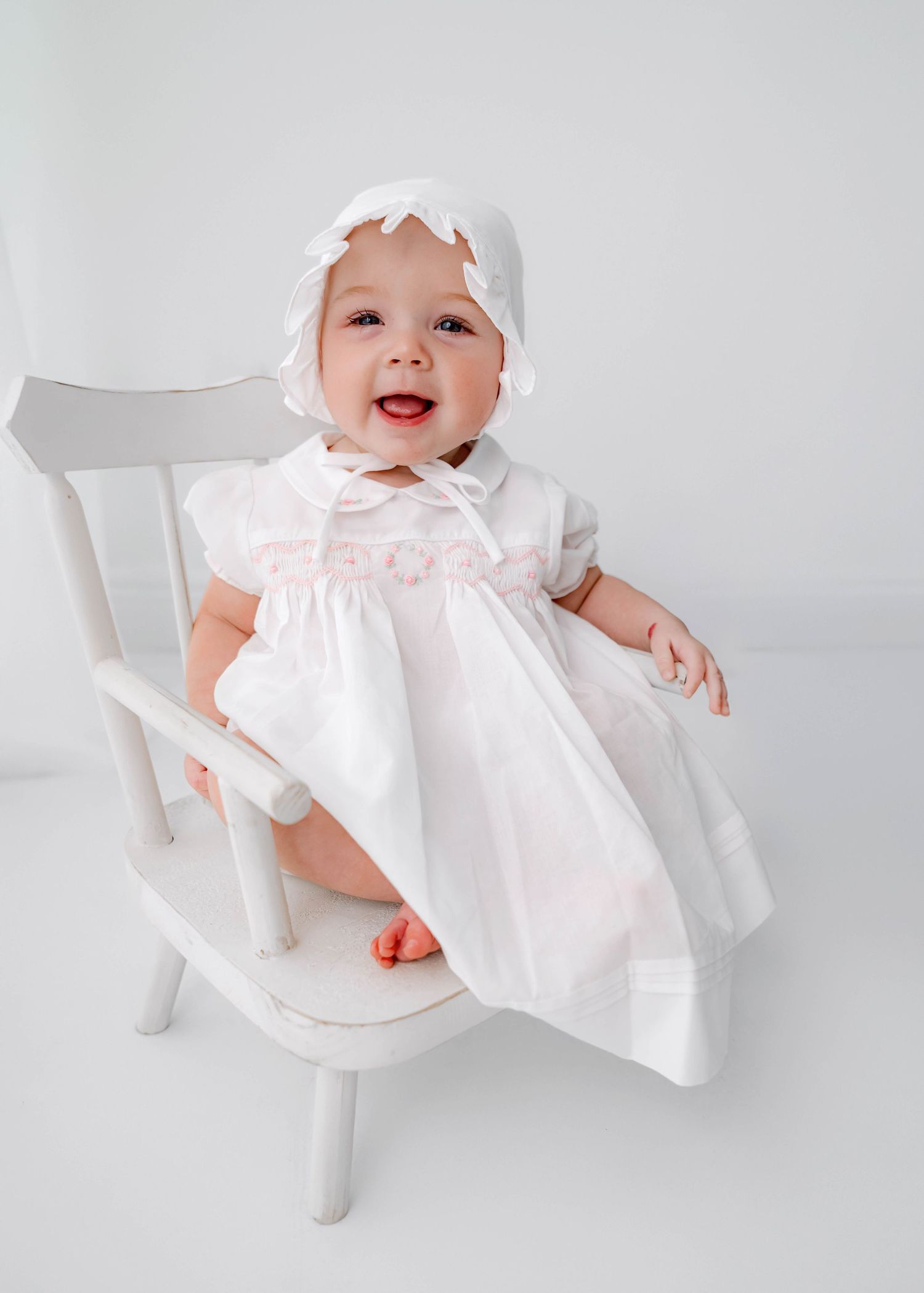 Smiling baby in white christening dress and bonnet sitting on a white wooden chair.