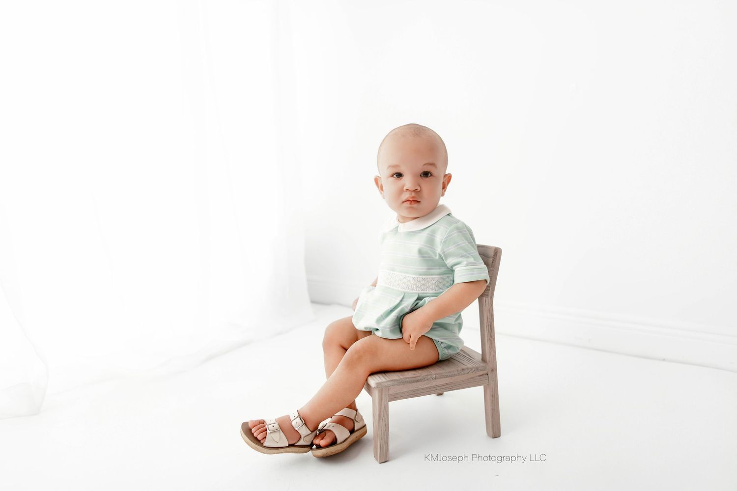 A baby boy sits on a small wooden chair in a bright white studio, wearing a mint romper and sandals.