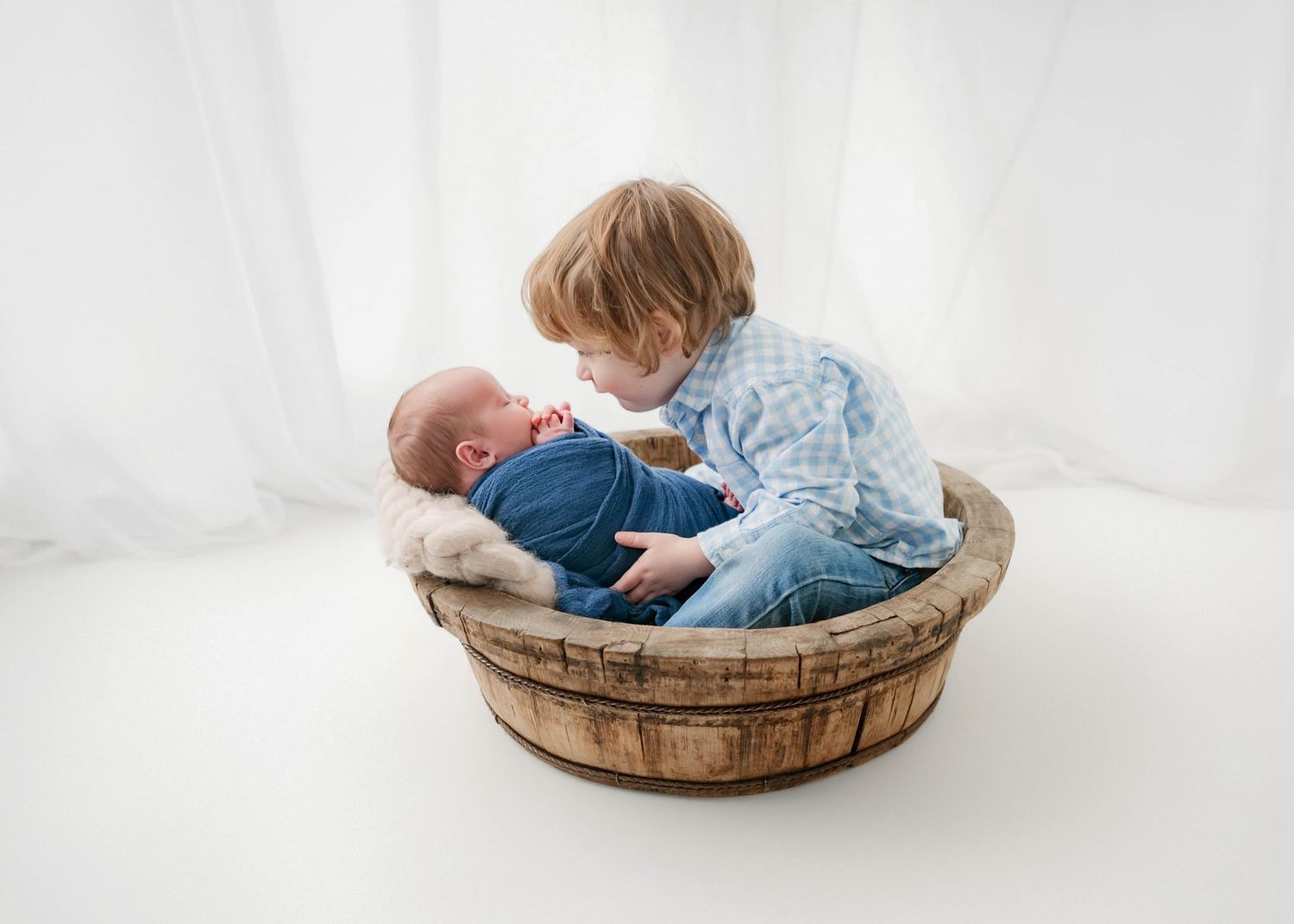 A toddler boy sits in a wooden barrel with a sleeping newborn baby wrapped in blue, sharing a tender sibling moment.