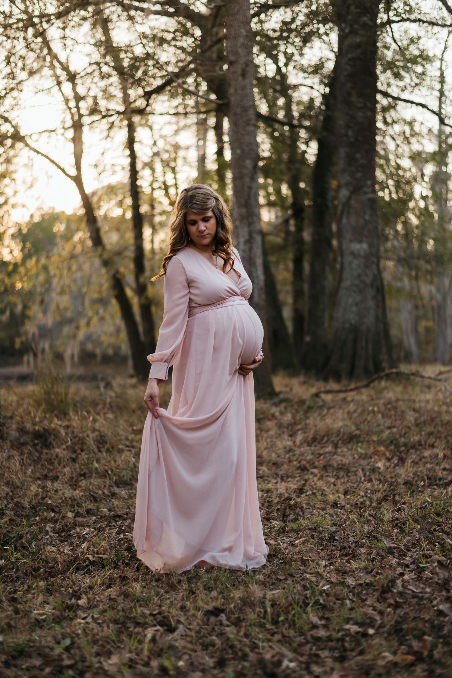 Woman in flowing pink dress poses gracefully among autumn leaves and tall trees at sunset.