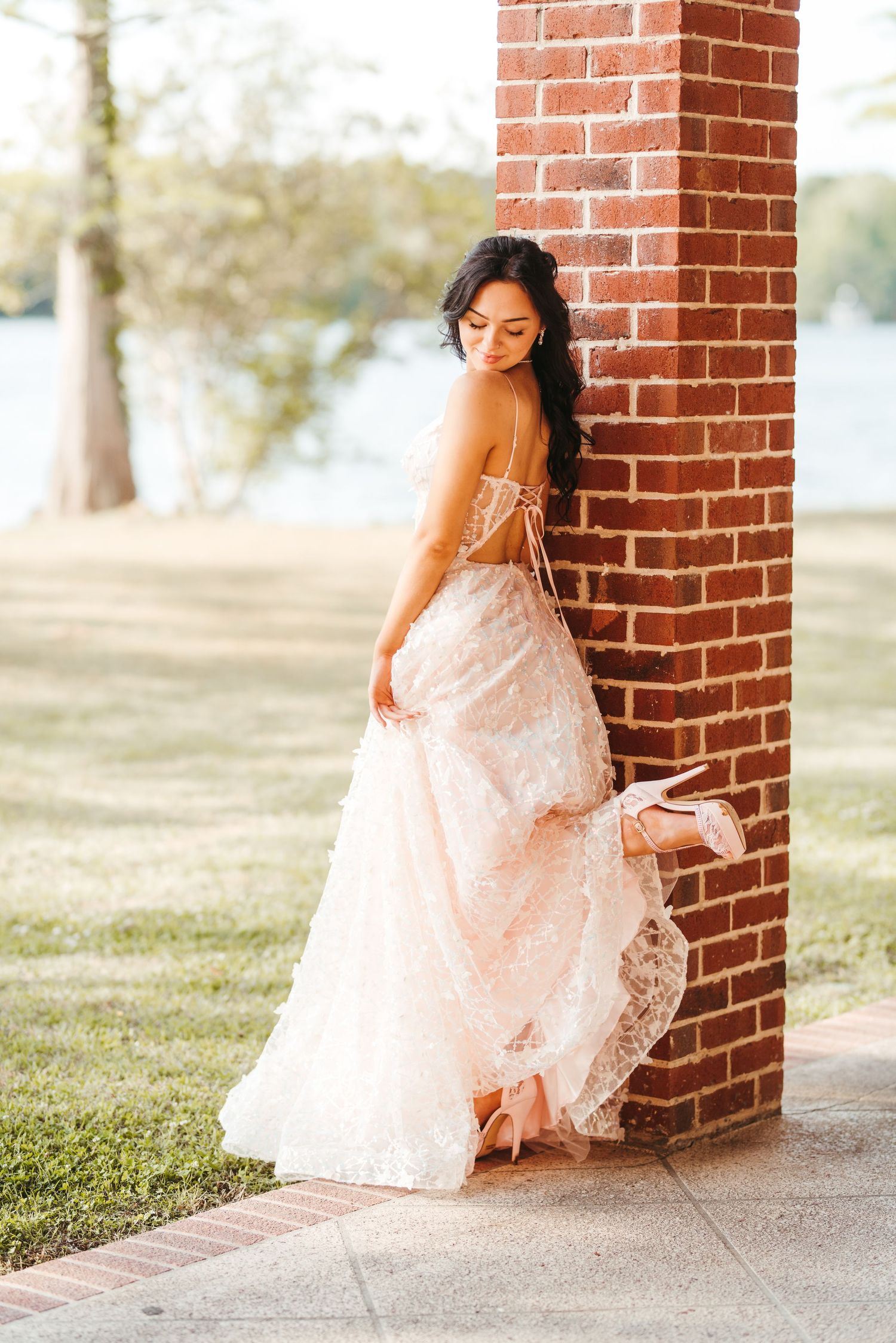 A romantic bridal portrait showing a flowing white lace gown against a brick column in an outdoor setting.