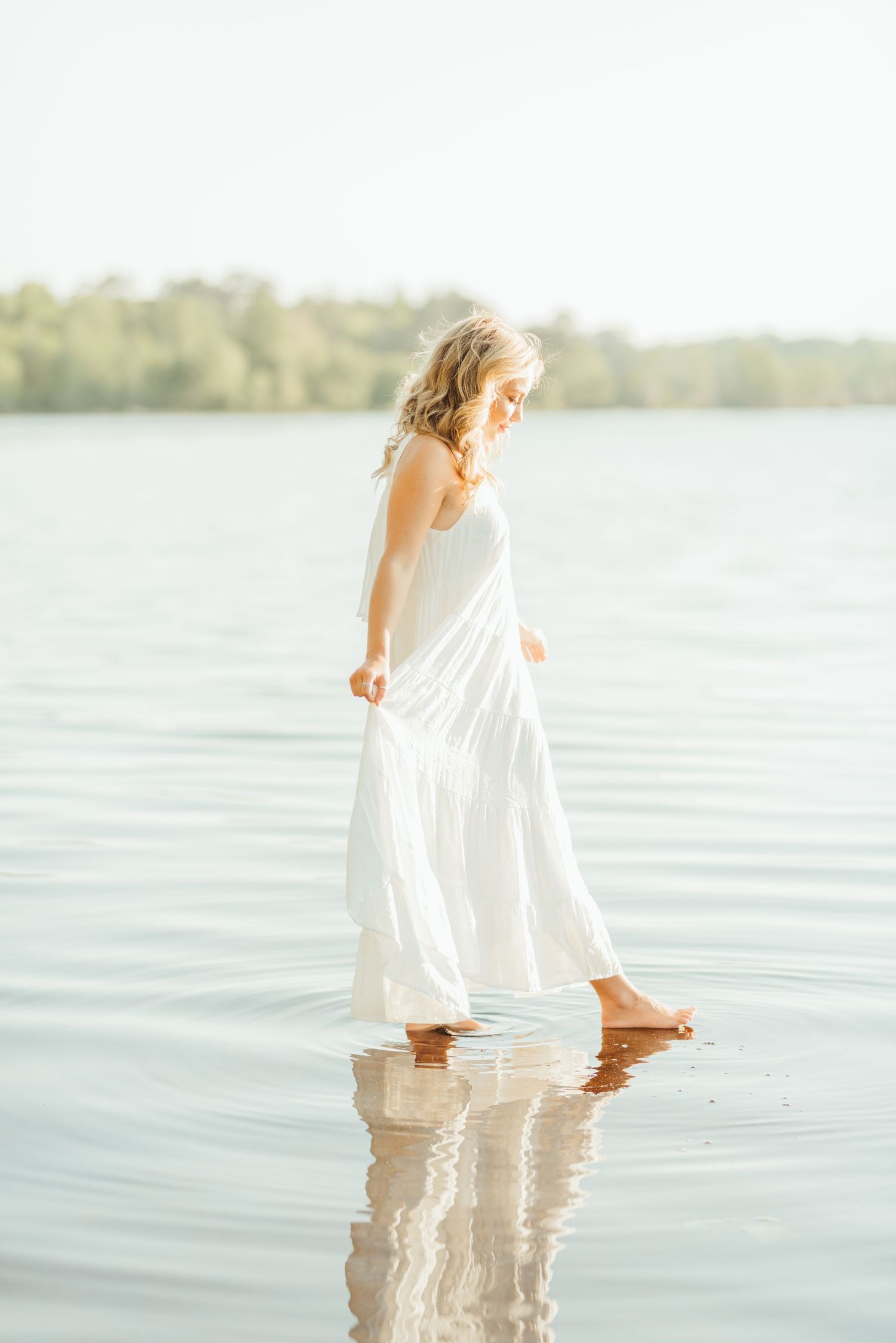 Person wearing white dress wading in shallow lake water during golden hour.
