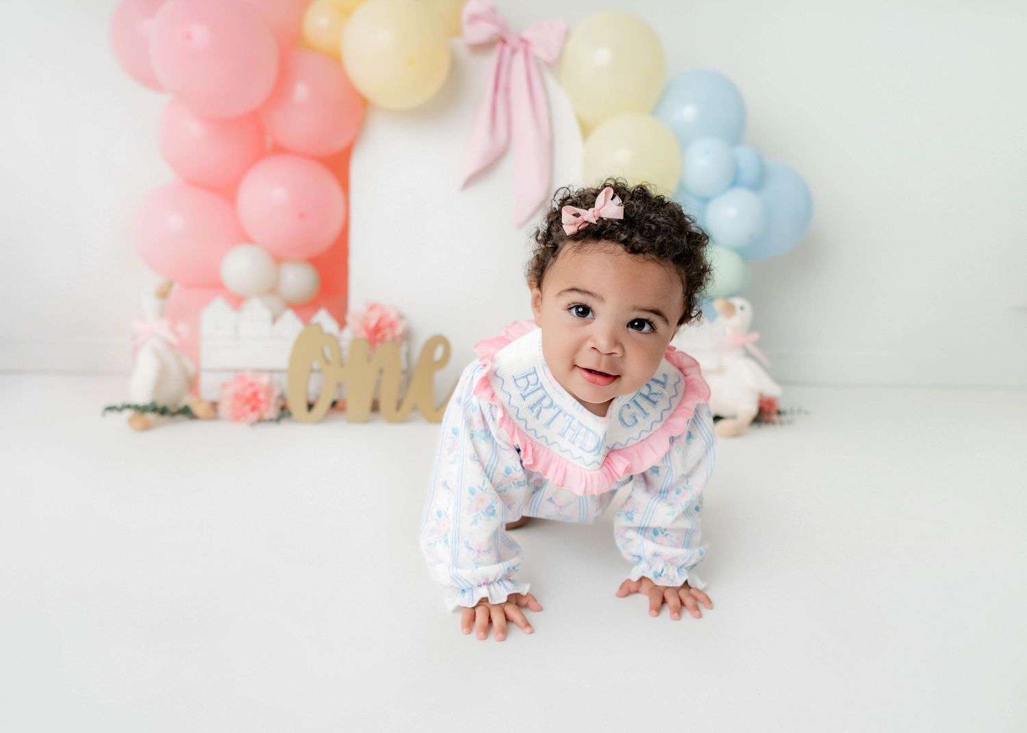 Baby girl in white outfit crawls on white floor with colorful balloon arch and gold crown for first birthday cake smash.