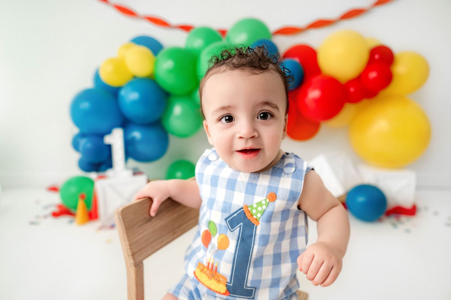 Baby boy in a number one outfit celebrates his first birthday with colorful balloons and a smash cake setup.