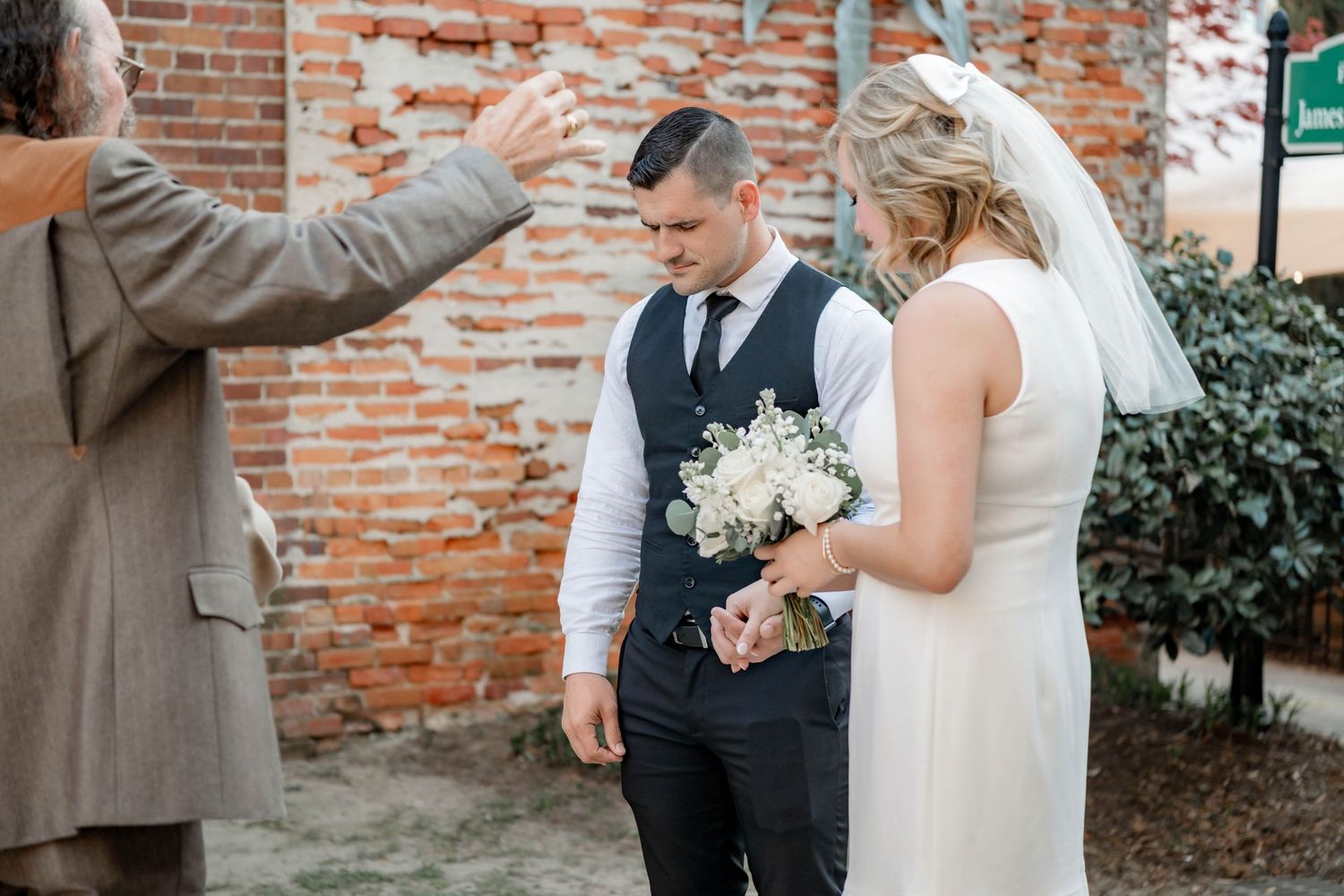 Bride and groom stand before officiant during outdoor wedding ceremony near rustic brick wall, bride holding white bouquet.