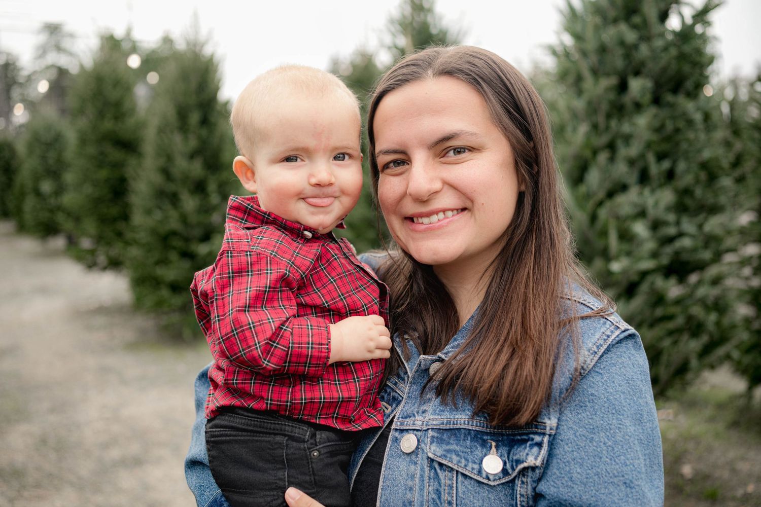 Young woman holding a smiling baby at a Christmas tree farm, both wearing casual winter outfits outdoors.