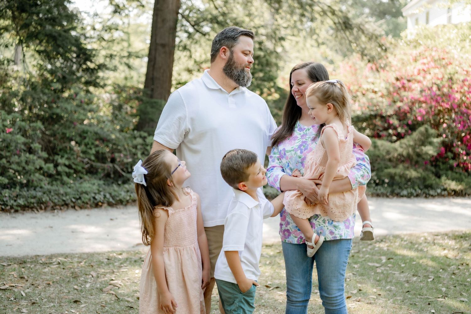 Family of five smiling together on a sunny park path surrounded by lush green and pink flowering trees.