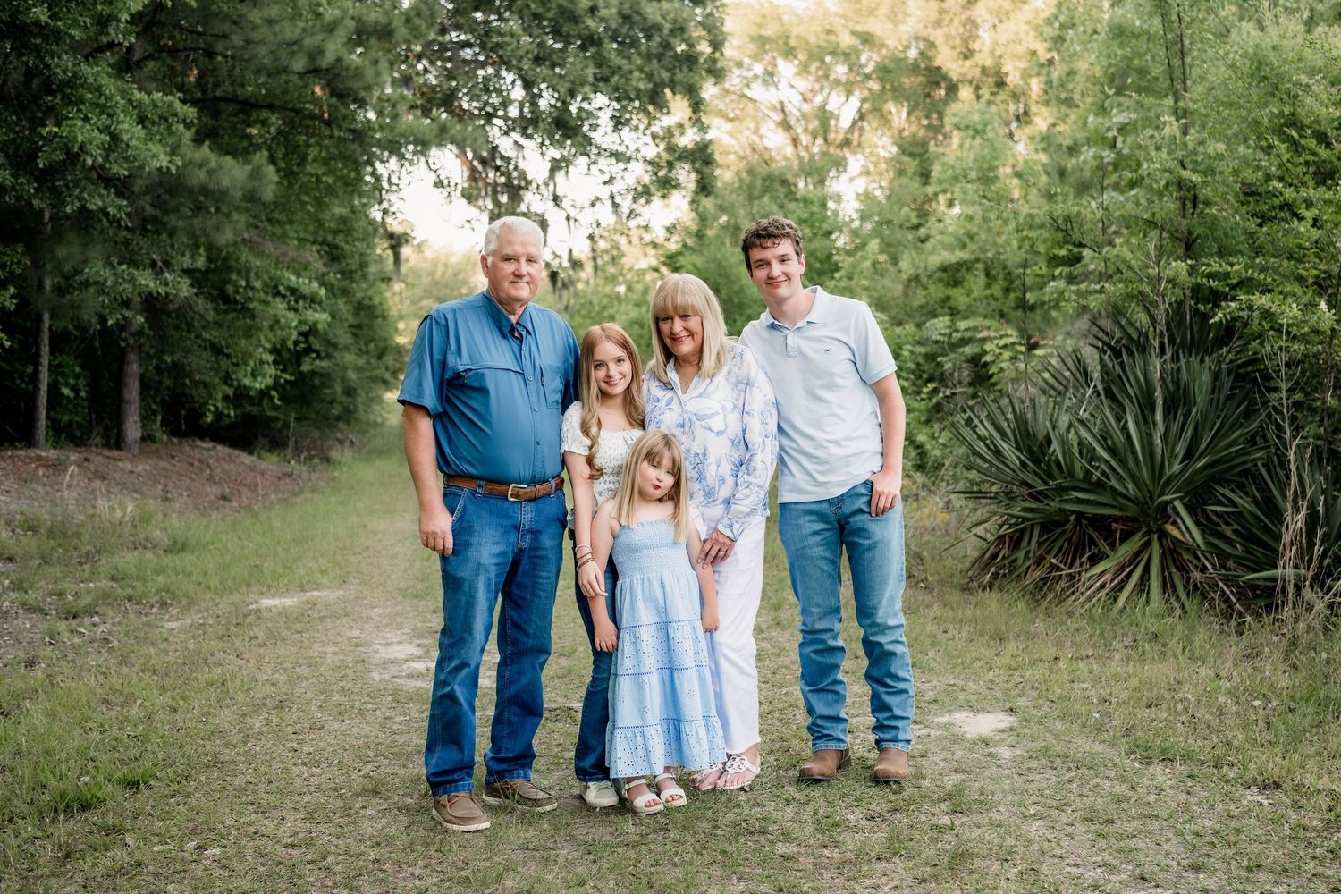 Family of five posing outdoors on a dirt path surrounded by lush green trees and vegetation.