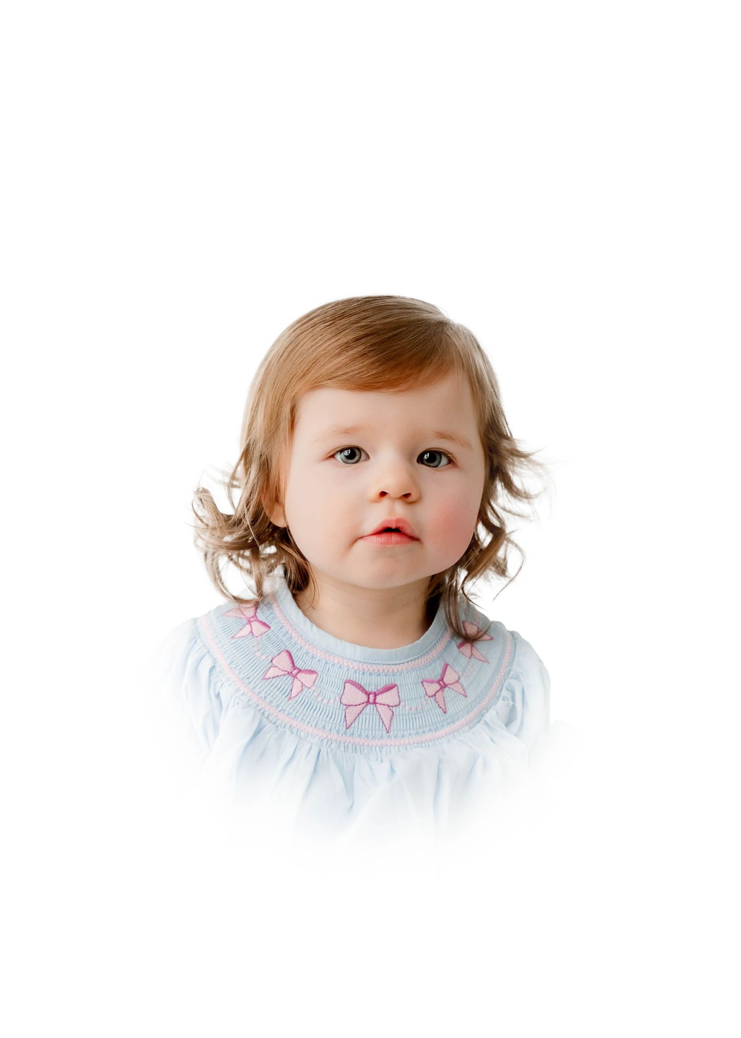 Toddler girl in a light blue smocked dress with pink bow embroidery, with curly brown hair, on white background.
