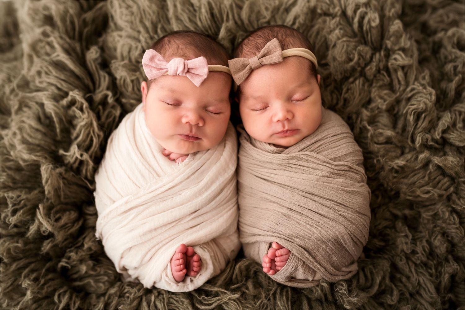 Twin newborns swaddled in cream wraps sleep peacefully side by side on textured blanket.