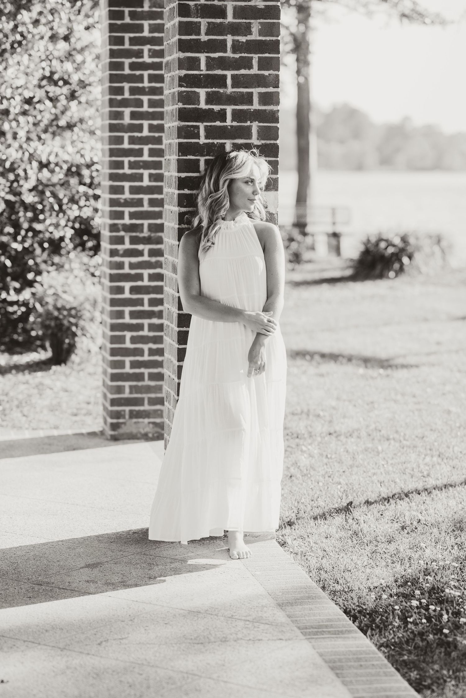 A graduate in a white maxi dress stands by a brick column in black and white.