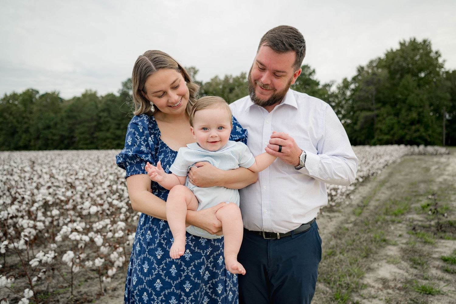 A young family poses in a cotton field, mother in blue floral dress and father in white shirt holding their baby boy.