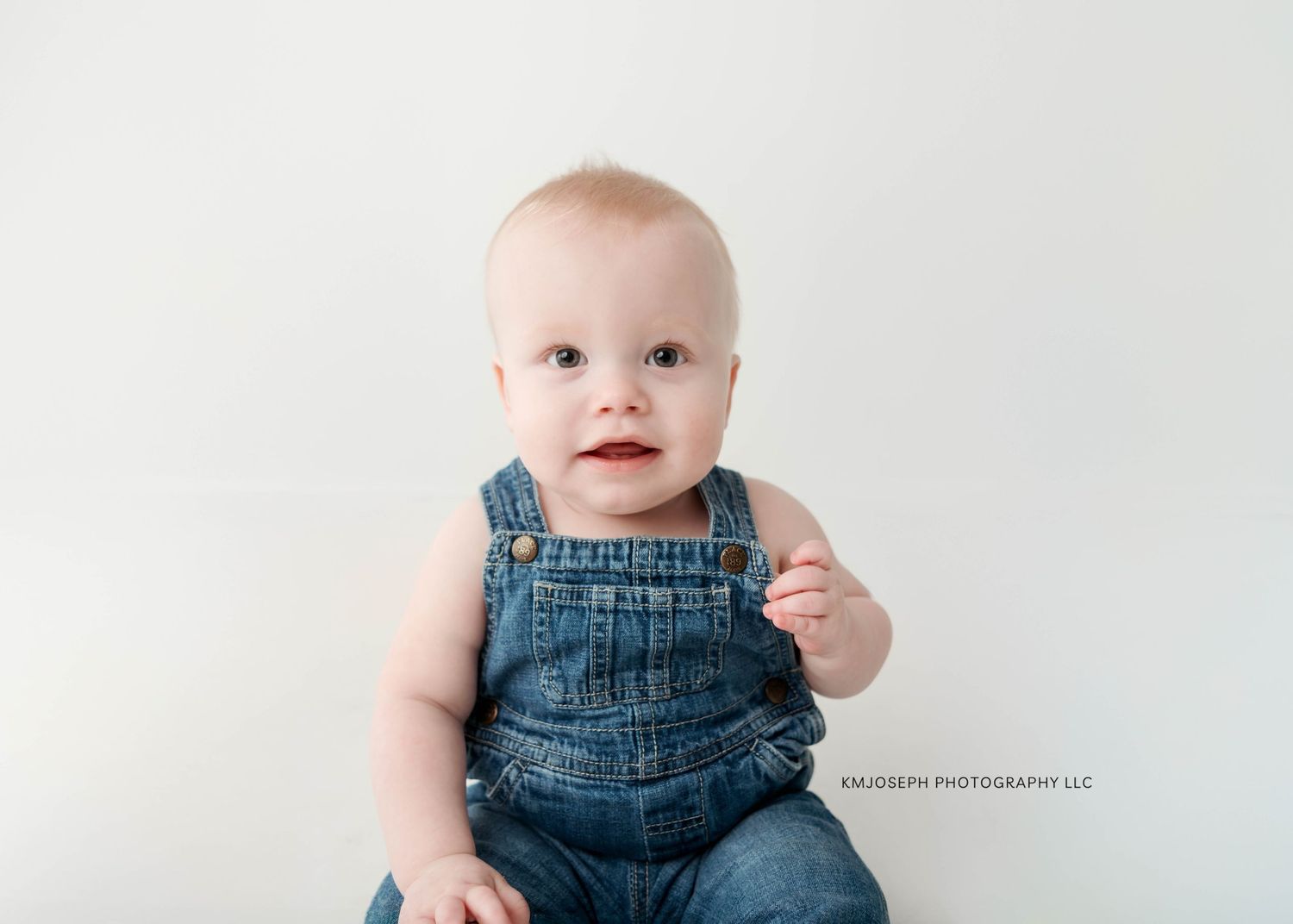 Baby in denim overalls sitting against white studio backdrop.