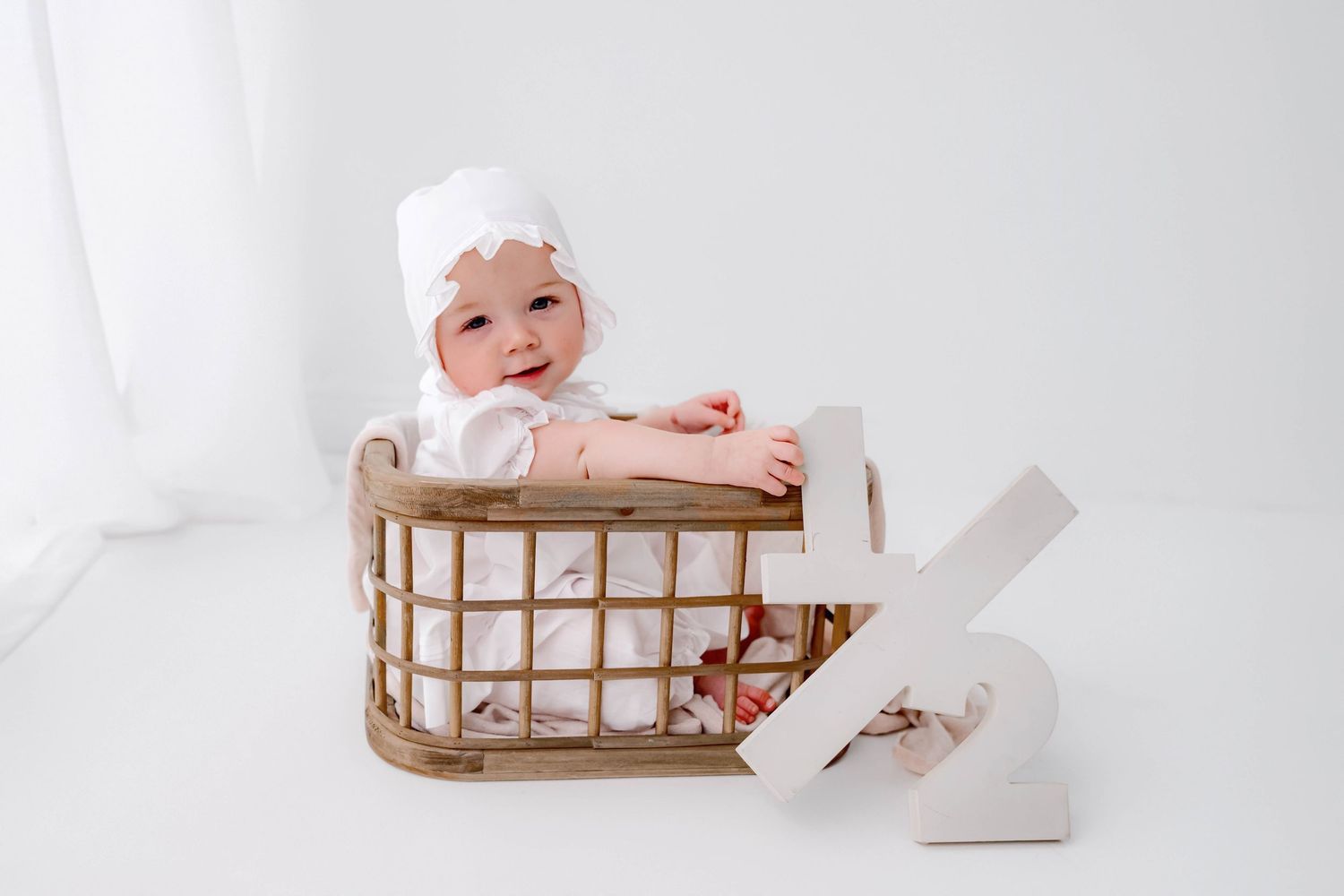 Smiling baby in white bonnet sitting in a wicker basket with toilet paper rolls on white background.
