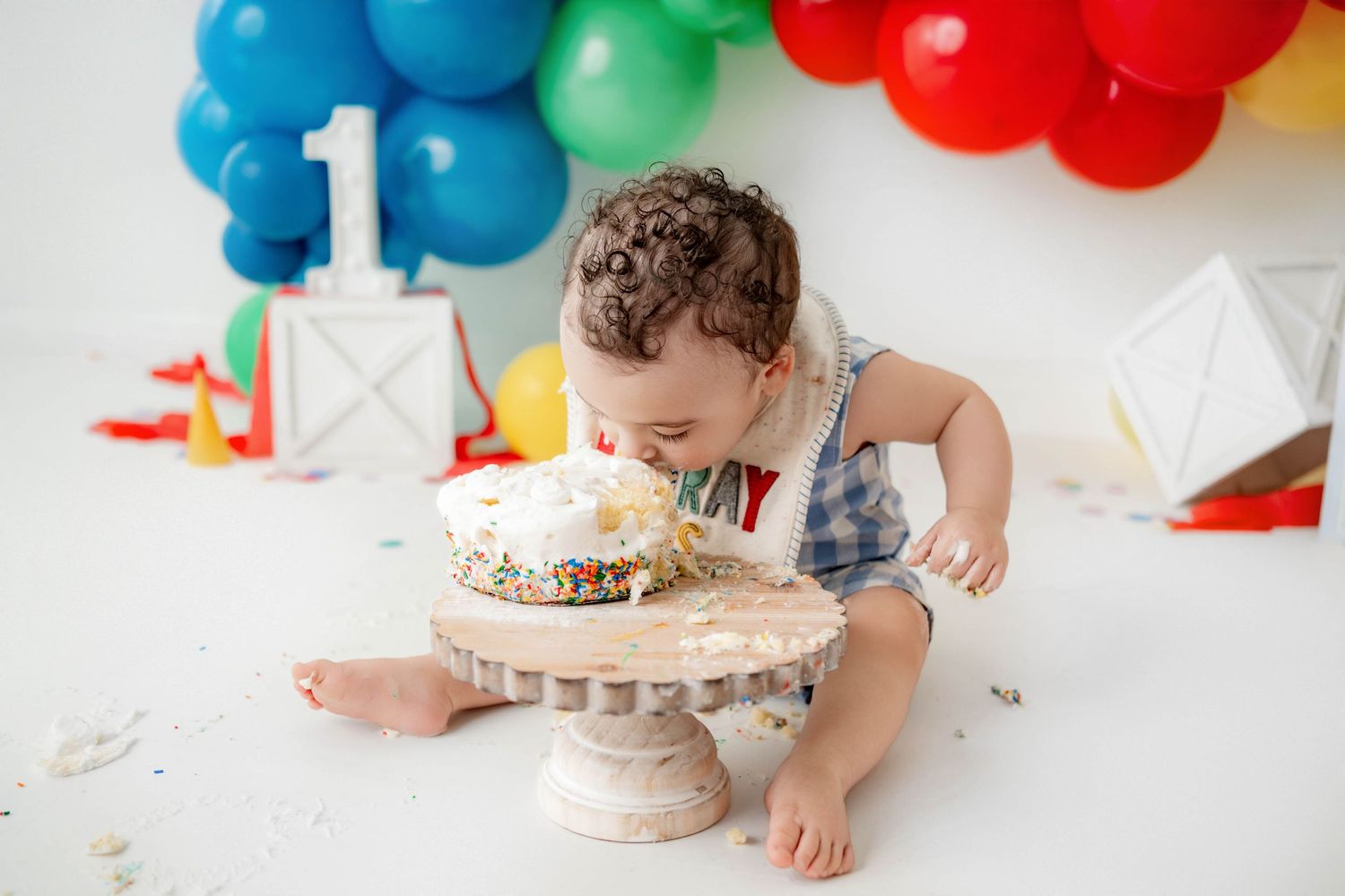Baby boy smashing his first birthday cake surrounded by colorful balloons and number one decorations.