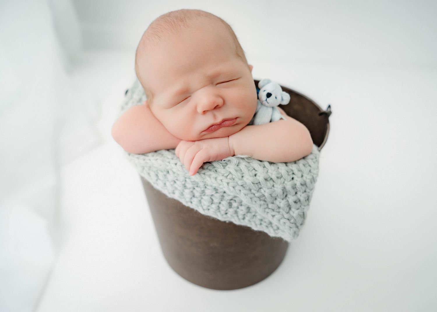 Sleeping newborn baby in a wooden bucket with gray knit blanket and tiny stuffed animal toy.