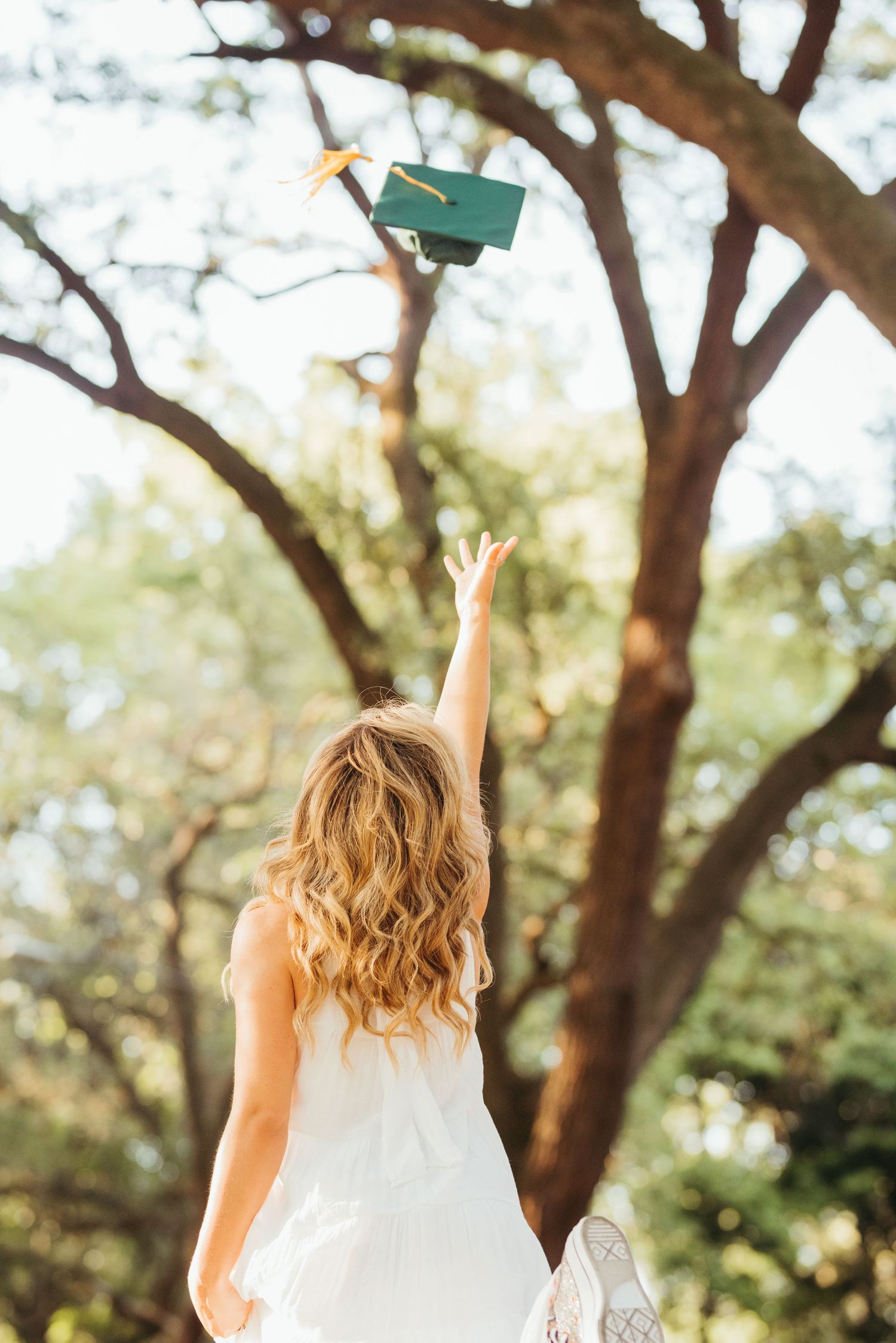 A graduate tosses a green graduation cap into the air beneath trees.