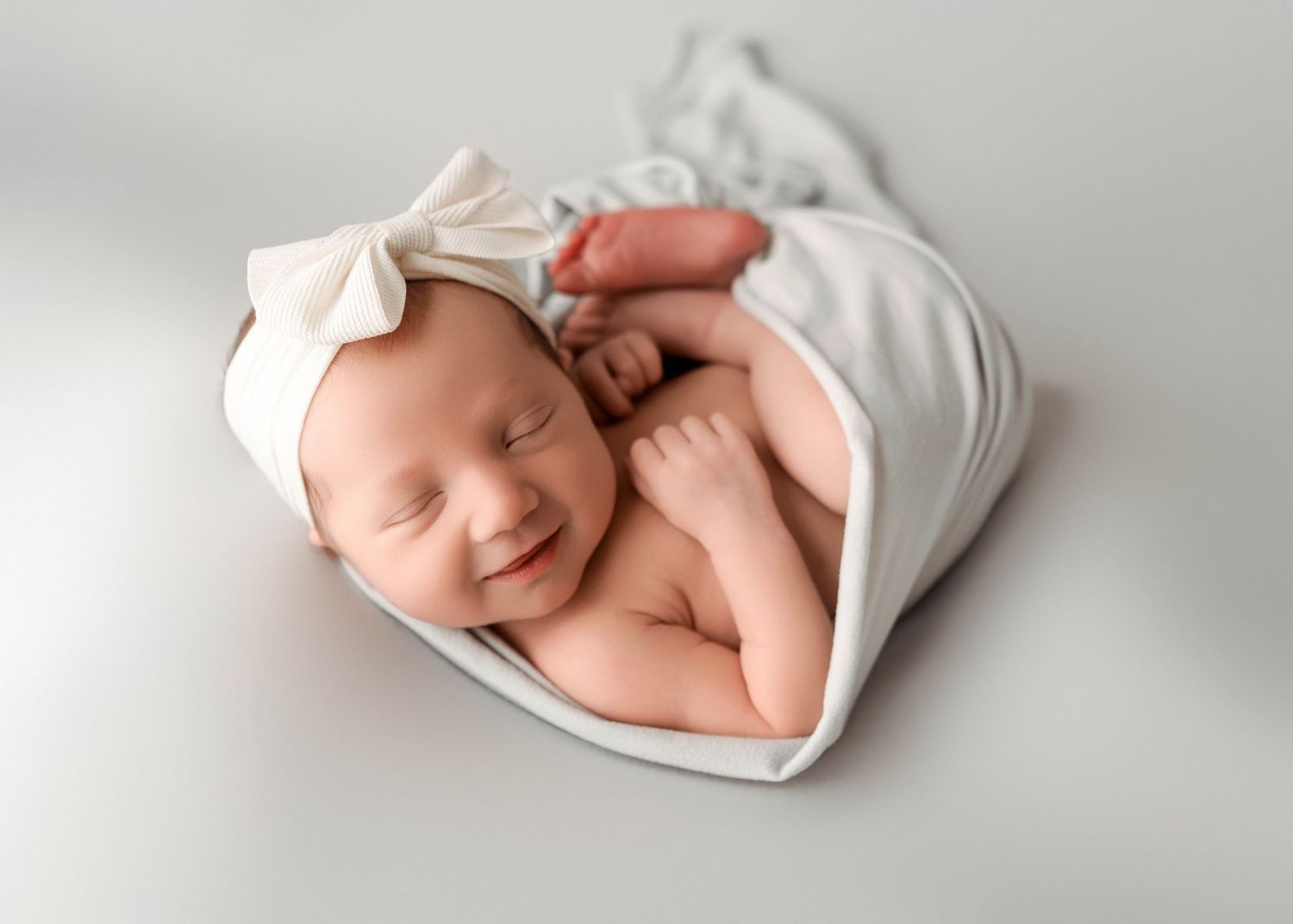 Sleeping newborn baby wrapped in white swaddle on neutral backdrop.