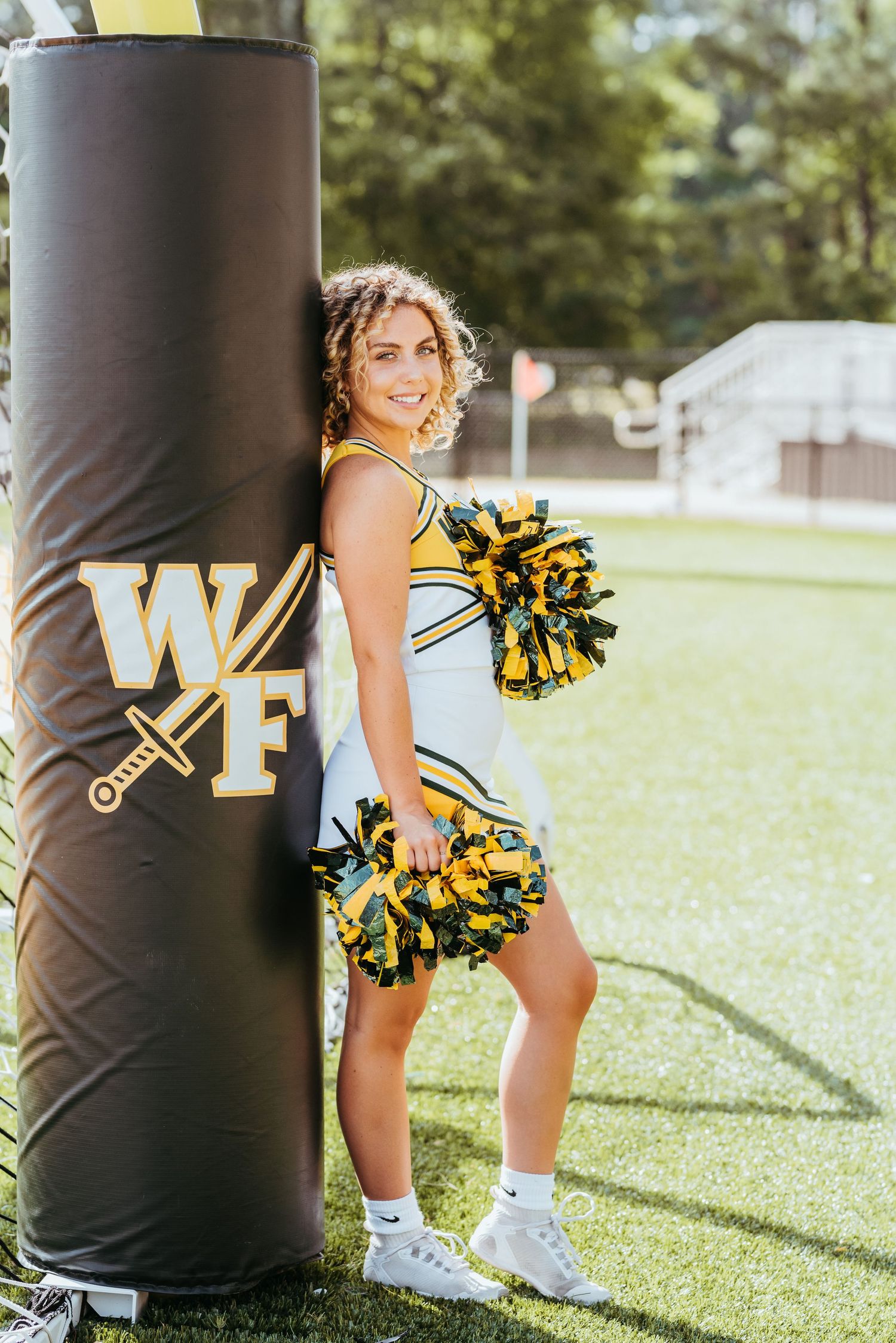 Cheerleader with pom poms poses next to black and gold football training equipment on grass field.