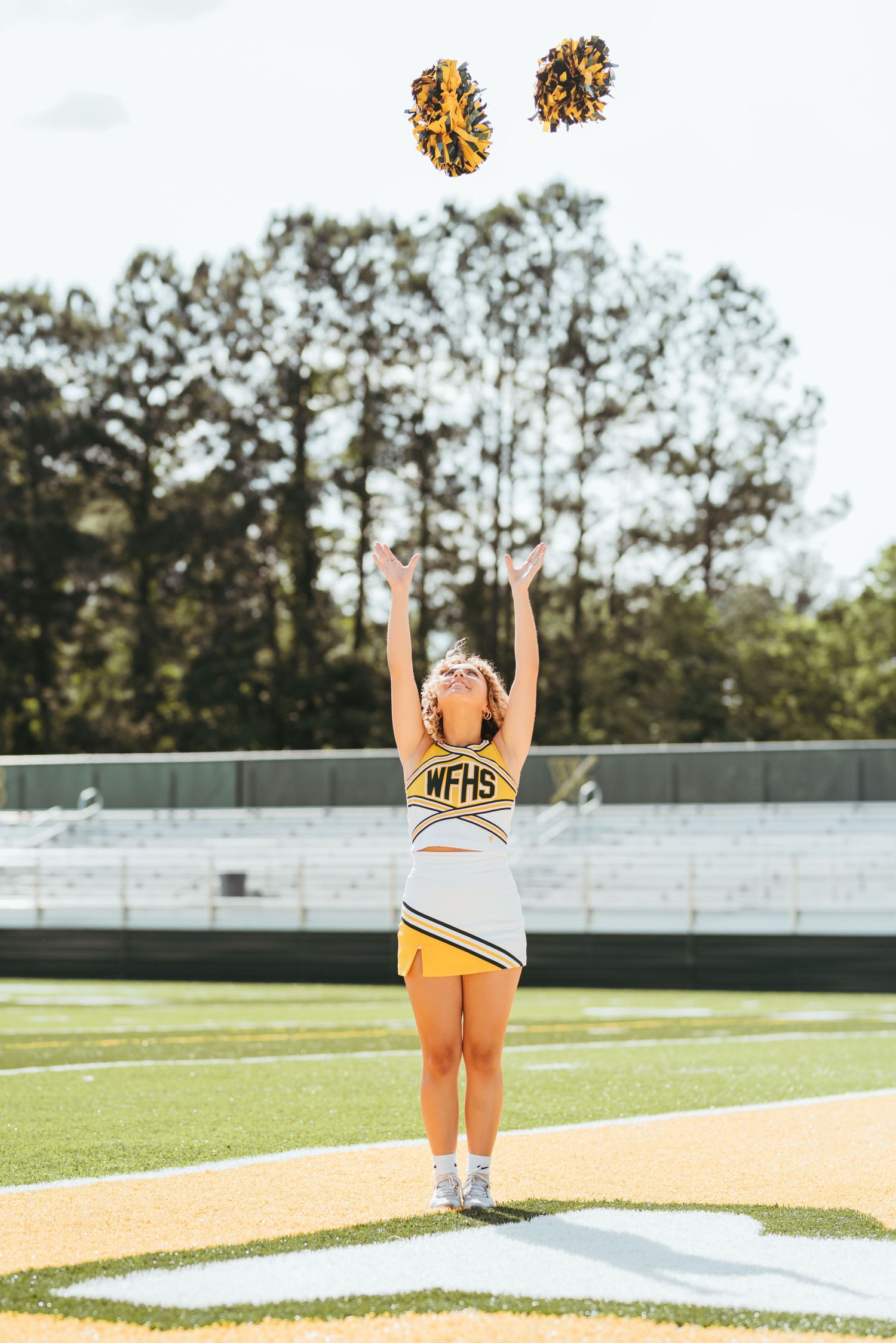 Cheerleader in yellow and black uniform poses on football field with raised arms against stadium bleachers.