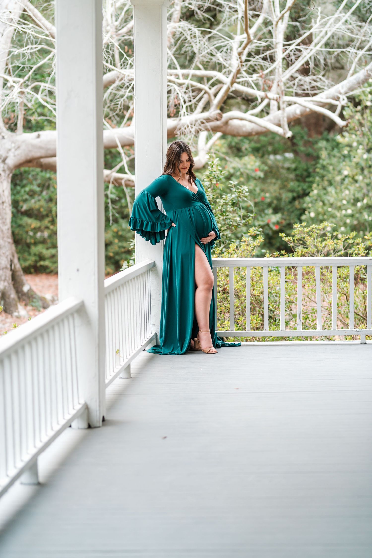 A couple poses together in a white gazebo surrounded by trees wearing emerald green and beige outfits.