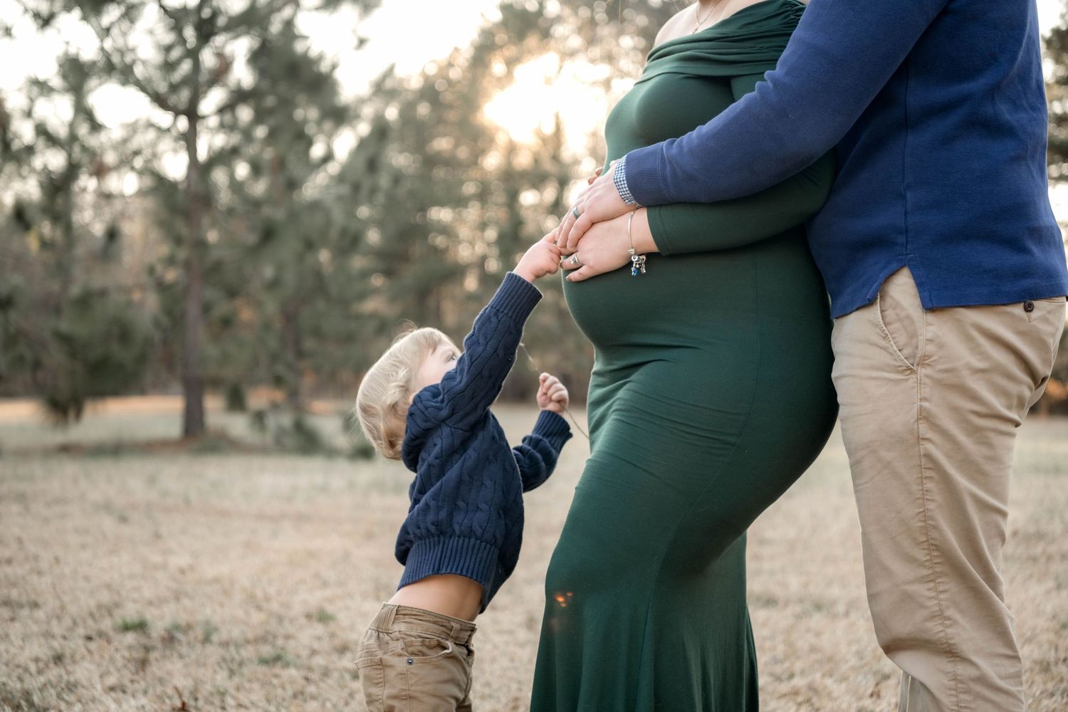 Pregnant woman in green dress with partner and toddler child holding hands outdoors in a park during golden hour.