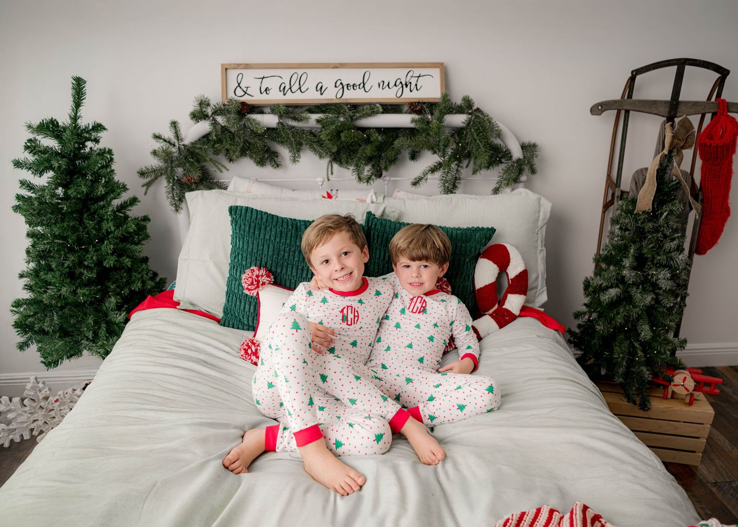 Two young children in matching Christmas pajamas sitting on a bed decorated with holiday greenery and festive decor.