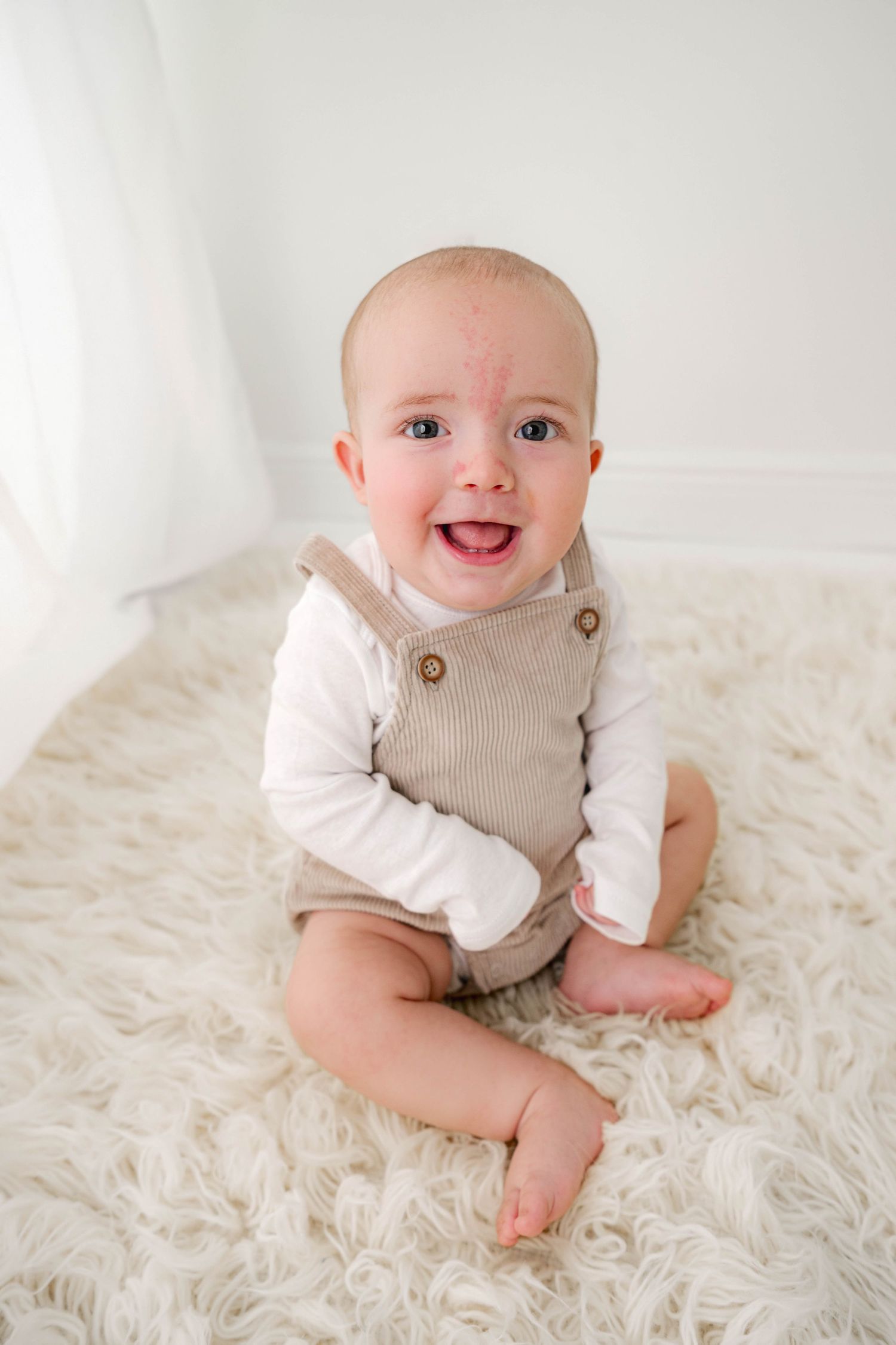 Smiling baby in beige overalls sitting on a fluffy white rug in a bright white room.