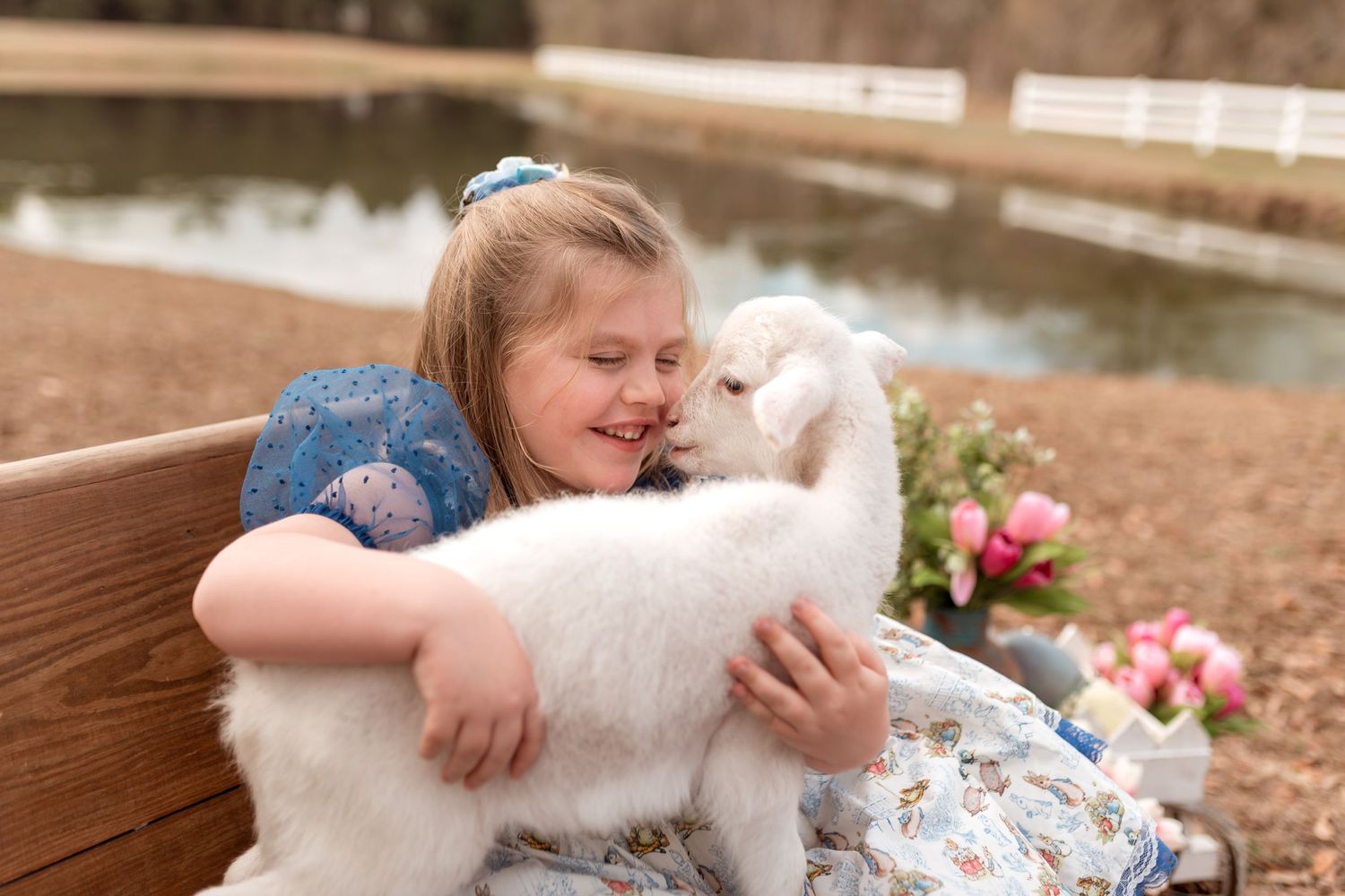 Young girl in blue dress hugging white fluffy lamb on wooden bench near pond.