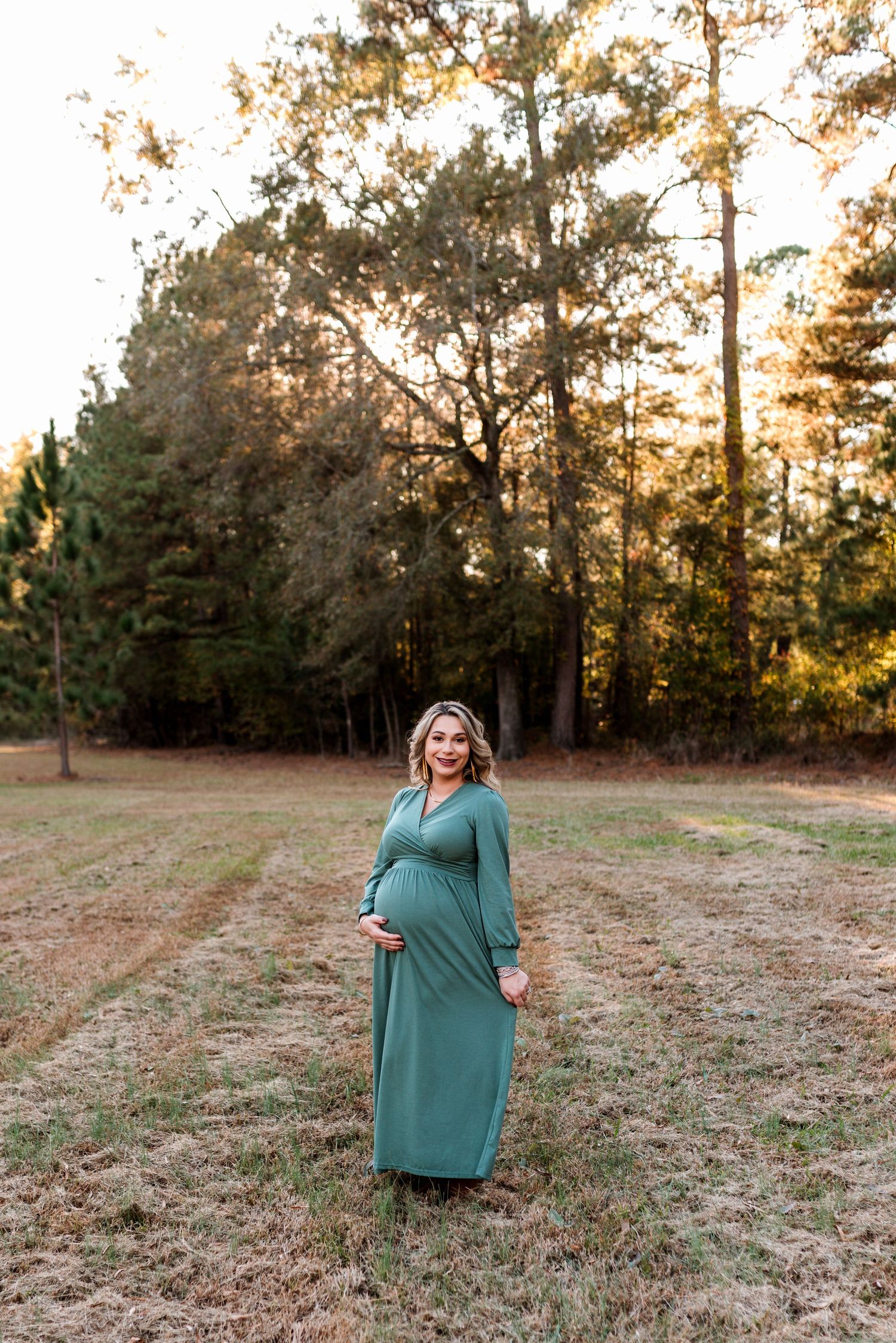 Woman in teal dress poses in golden sunlit field with pine trees.
