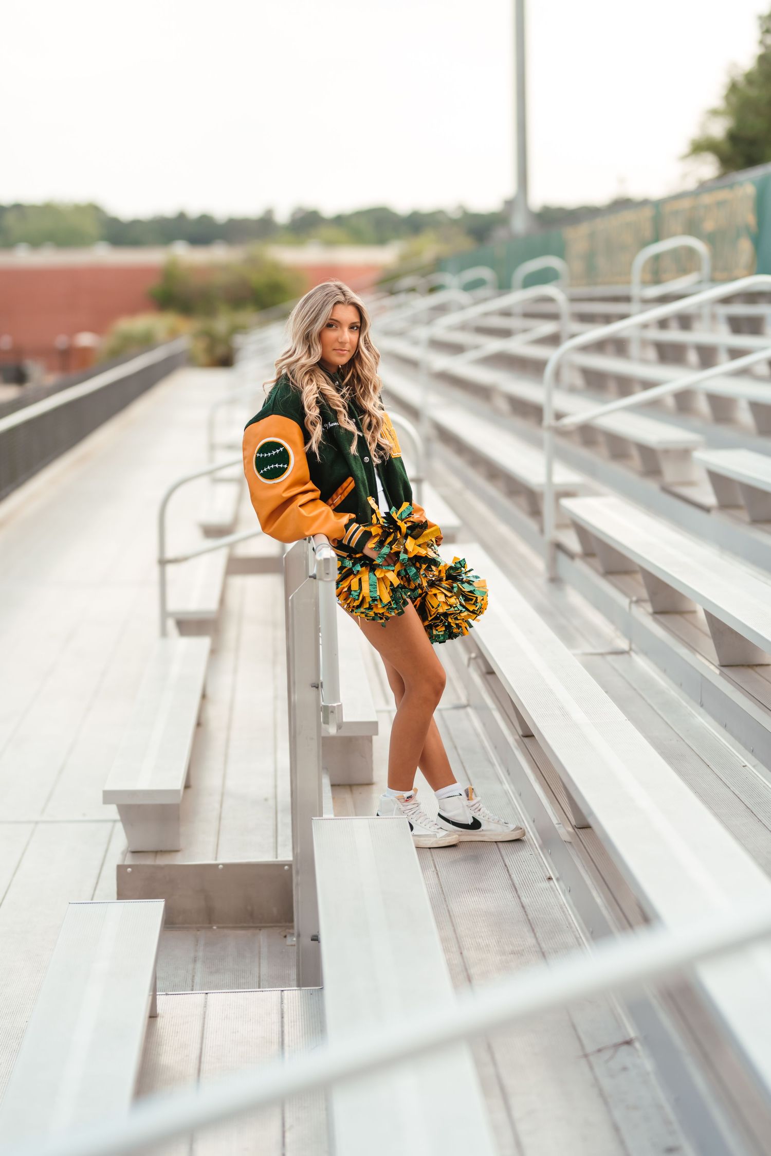 Cheerleader posing on stadium bleachers with pom poms and varsity jacket.