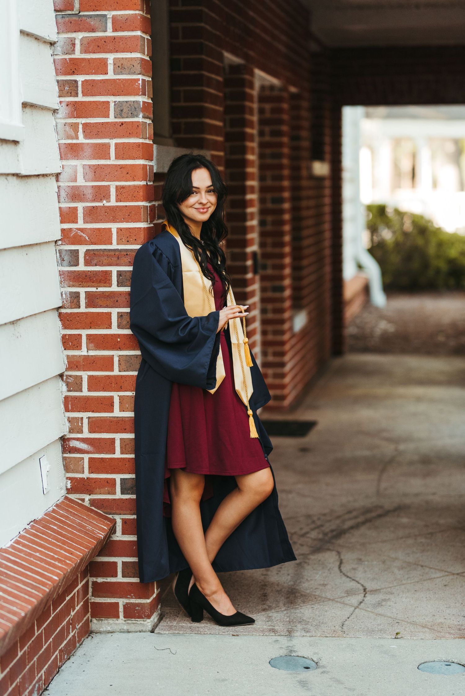 A graduate in a burgundy dress and navy gown poses casually against brick architecture.
