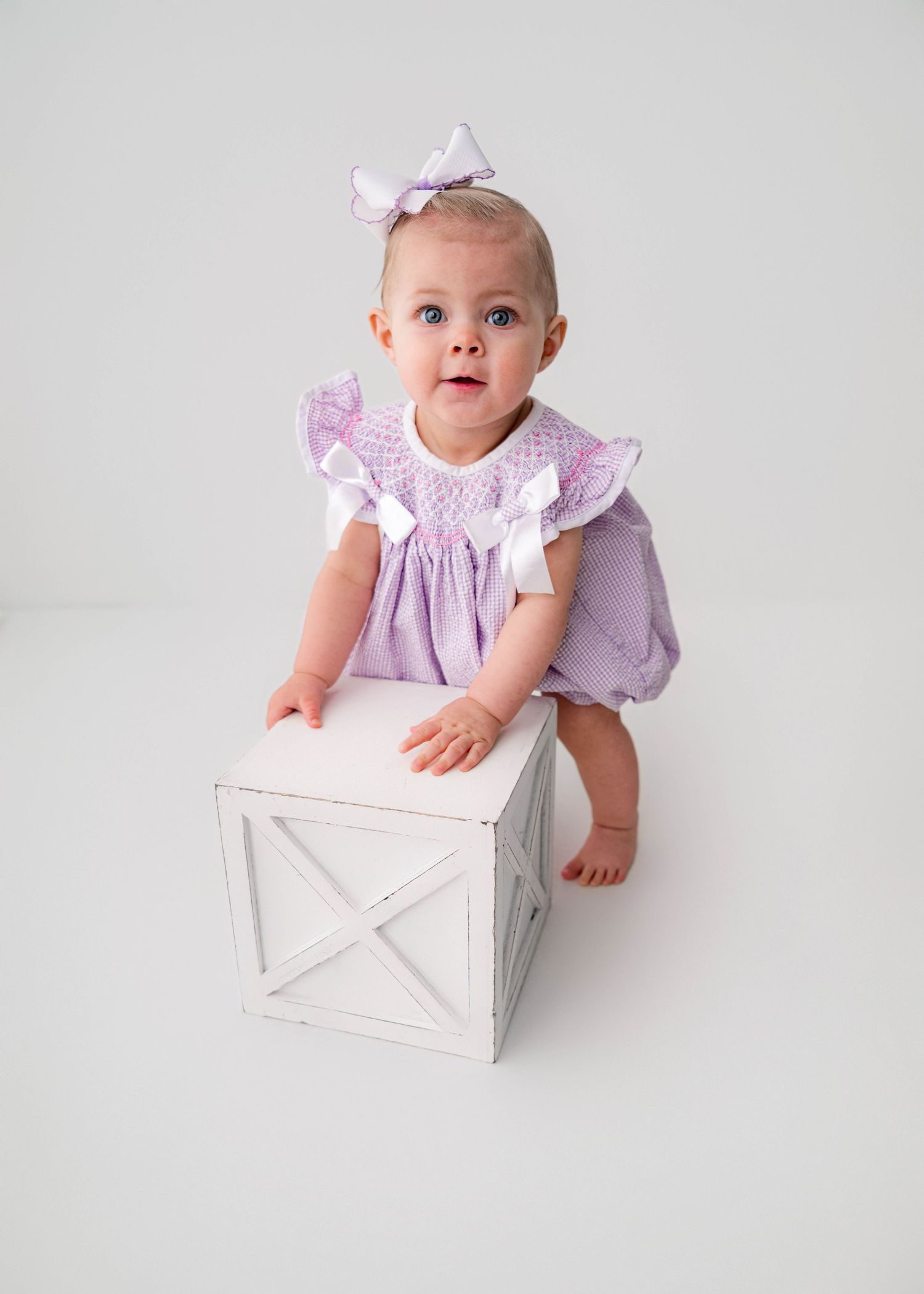 Baby girl in lavender ruffle romper with white bow headband poses by white wooden cube.