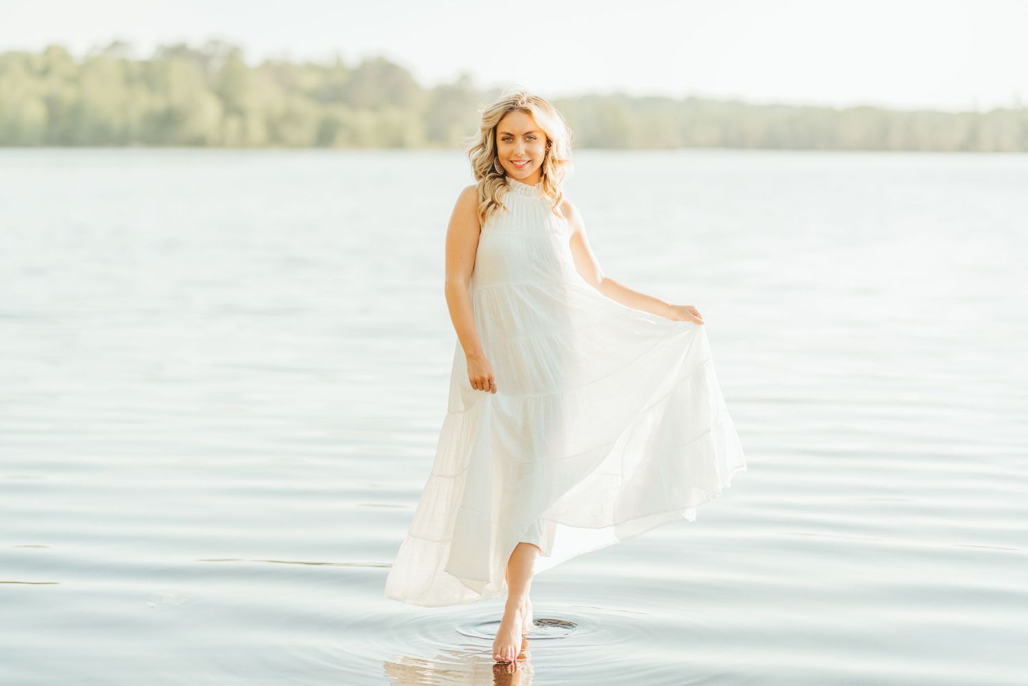 Person in flowing white dress walking along tranquil waterfront at sunset.