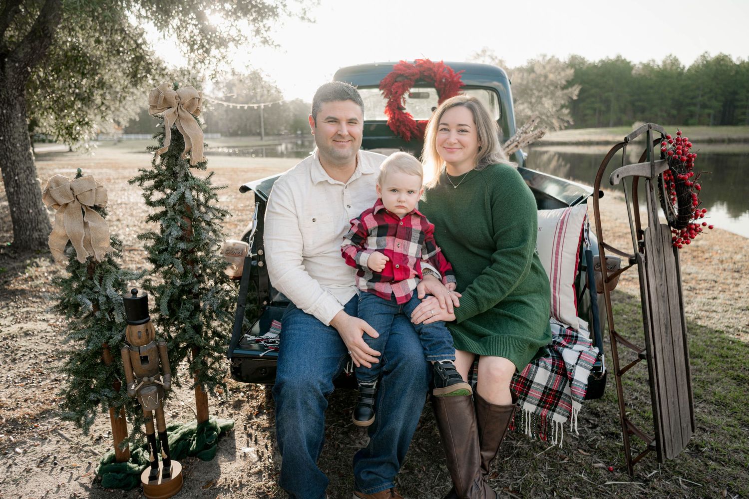 Family of three posing for a Christmas photo outdoors near a decorated vintage truck with holiday wreaths and trees.