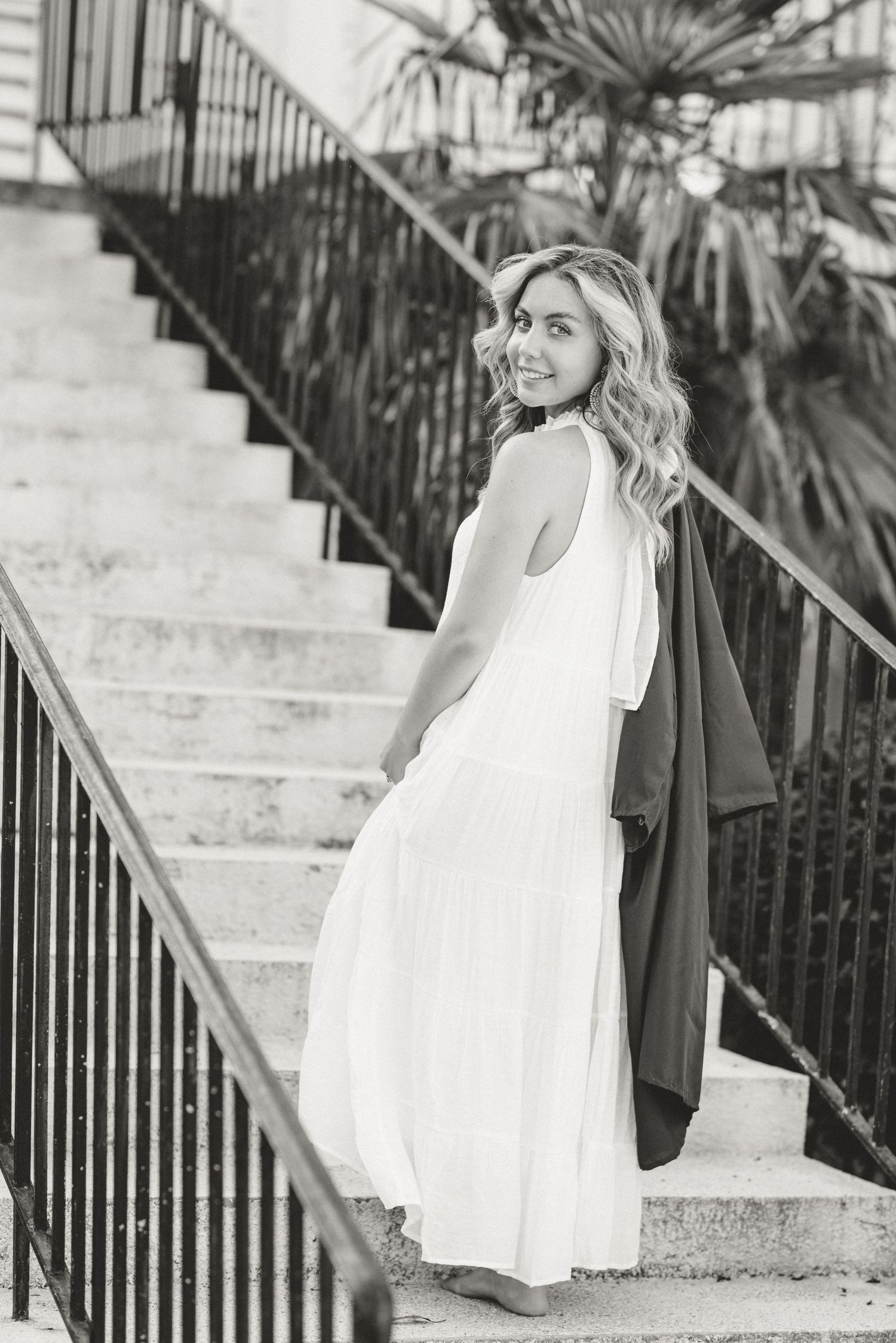 A graduate in a flowing white dress poses on stone steps in black and white.