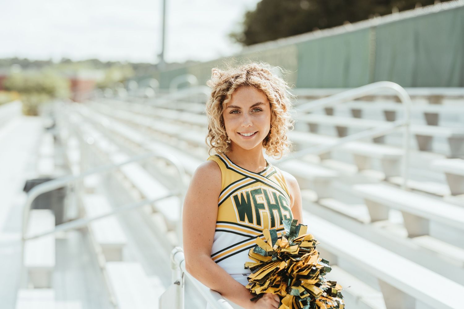 Cheerleader in yellow uniform posing on stadium bleachers with pom poms.