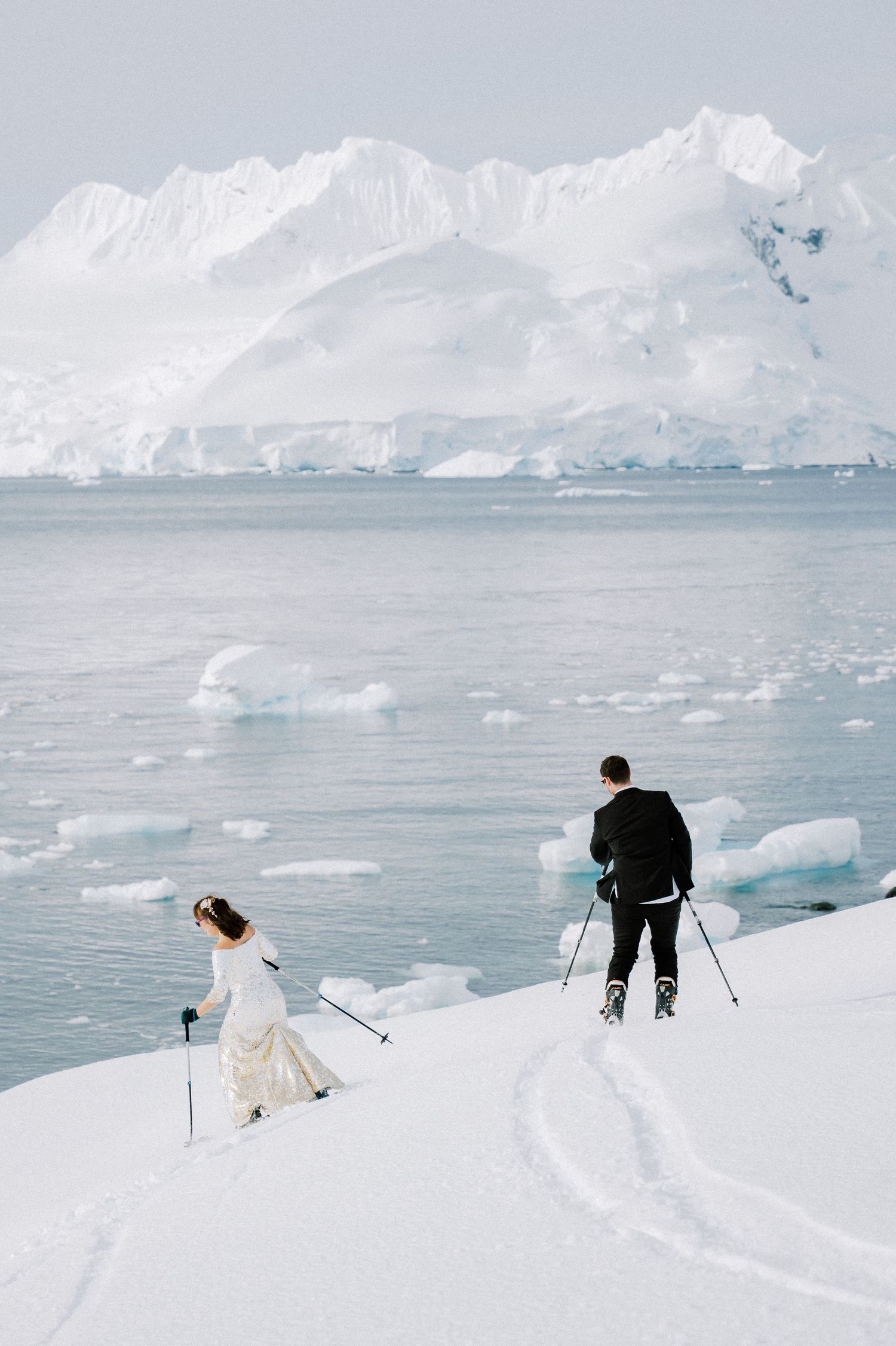 Two skiers descend a snowy slope with dramatic glacial landscapes and icebergs in the background.