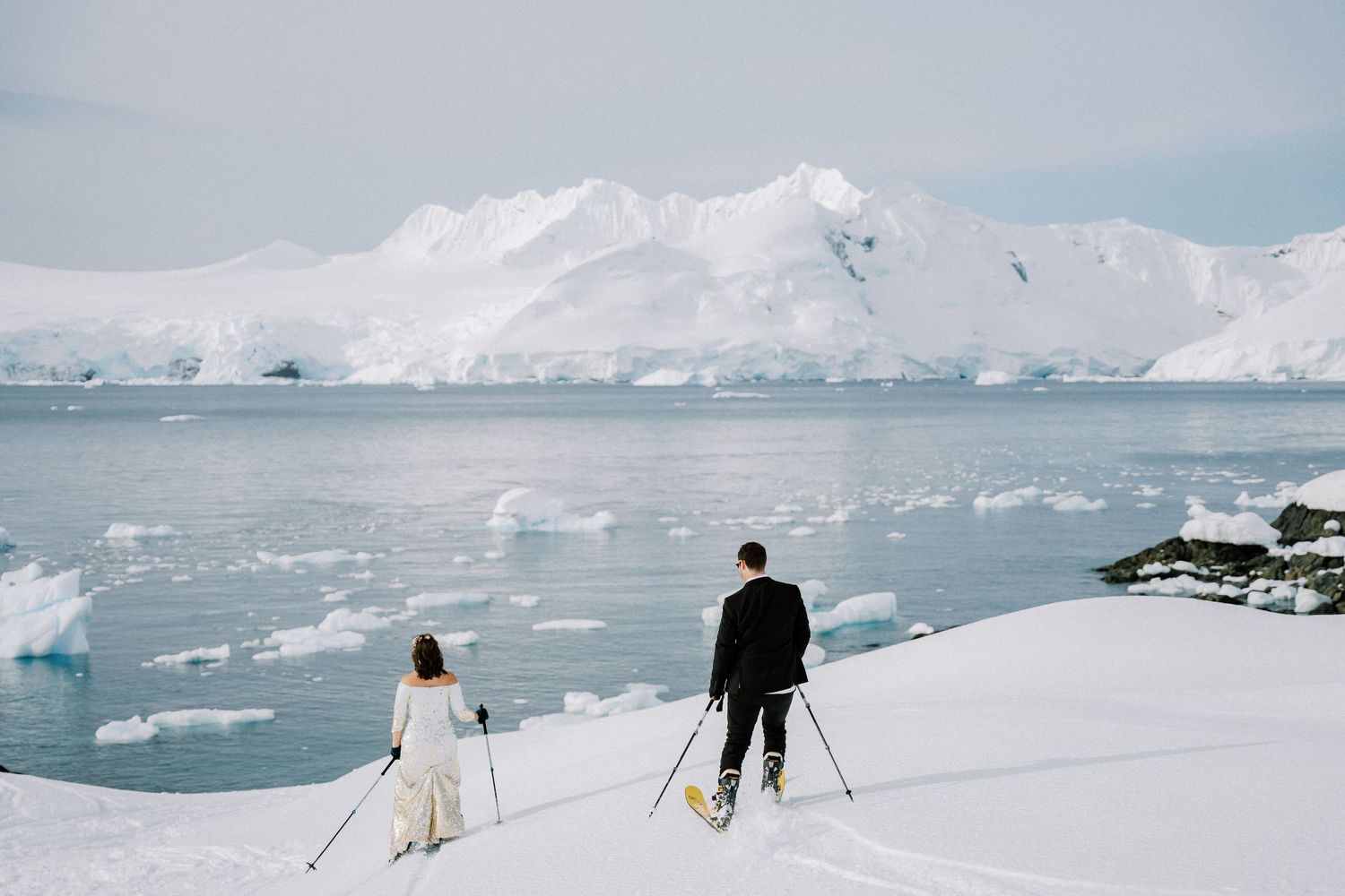 Two people ski together on snowy slopes with glacial mountains and icy waters in the distance.