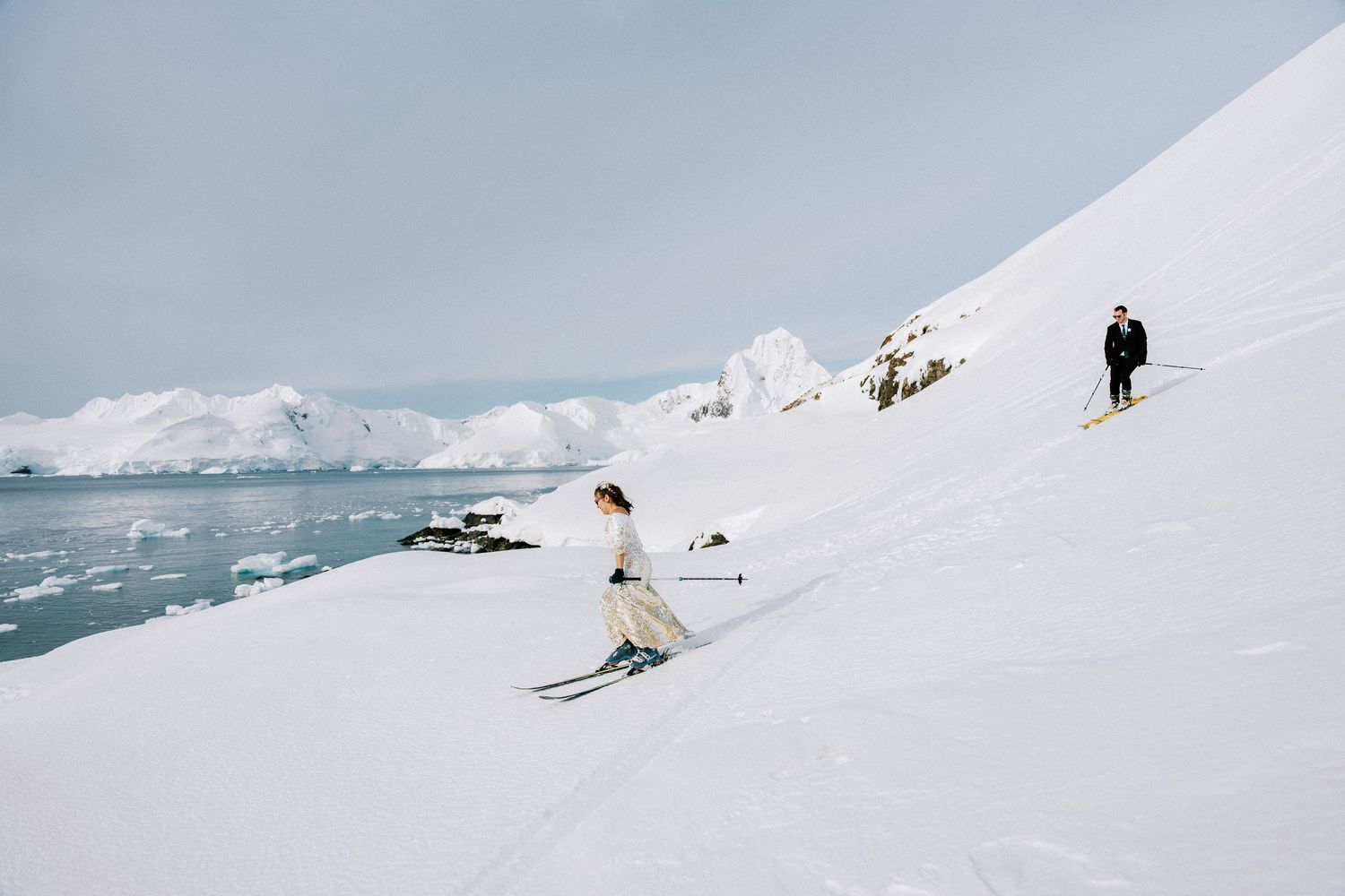 Skiers navigate pristine snow slopes alongside an icy coastline in a dramatic polar landscape.